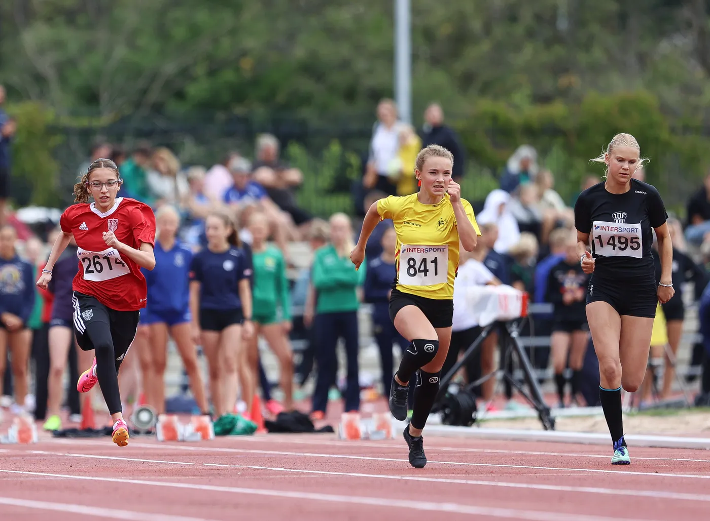 تظهر ثلاث عداءات يسجلن أوقاتاً رائعة في سباق، حيث ترتدي العداءة في المقدمة قميصاً أصفر وتبدو متفوقة، بينما العداءة على اليسار ترتدي قميصاً أحمر. يوجد جمهورٌ يتفرج في الخلفية، مما يضيف جوًا حيويًا للمنافسة.