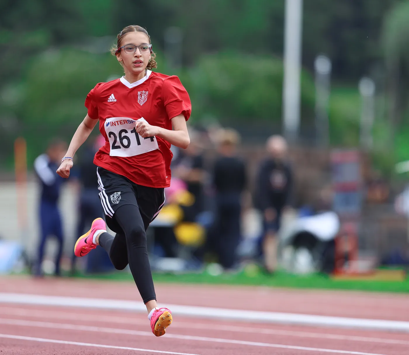 عداءة شابة ترتدي قميصًا أحمر وسروالًا أسود، تحمل رقم 2614، وهي تجري بسرعة على حلبة سباق رياضي. يُظهر خلفها مجموعة من الأشخاص في حالة حركة، مع منظر طبيعي أخضر في الخلفية.