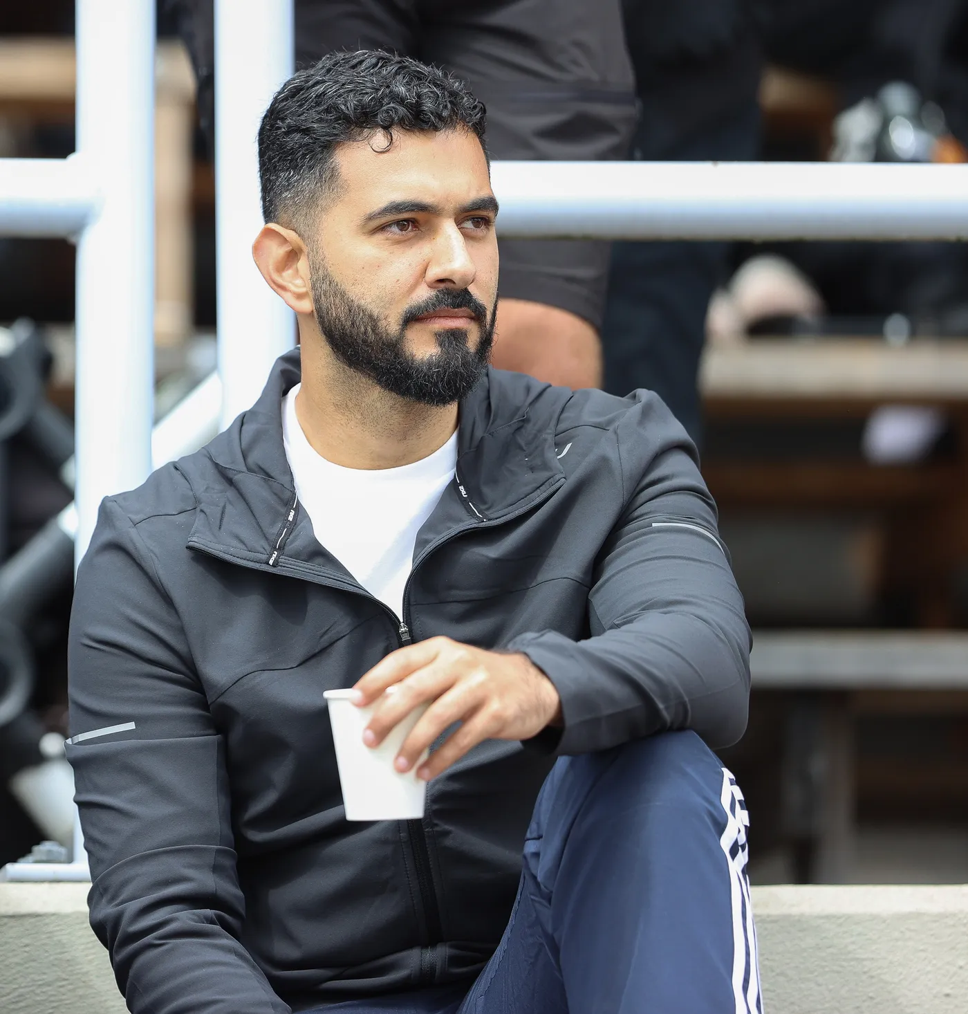 رجل يجلس في منطقة مخصصة للمشاهدين، يرتدي سترة رياضية داكنة وسروالاً رياضياً. يحمل كوباً ورقياً بيده، وينظر بتركيز نحو شيء أمامه.