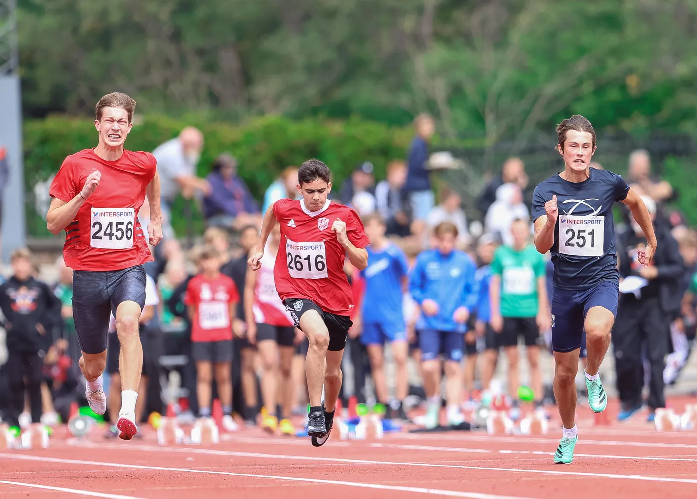 يظهر ثلاثة عدائين يتنافسون في سباق على مضمار رياضي، يرتدي اثنان منهم قمصان حمراء ويظهرون بشكل متحمس أثناء الجري، بينما يرتدي الثالث قميصًا أسود. الخلفية مليئة بجماهير ومشجعين يرتدون ملابس متنوعة، مما يضيف أجواء حماسية للحدث.