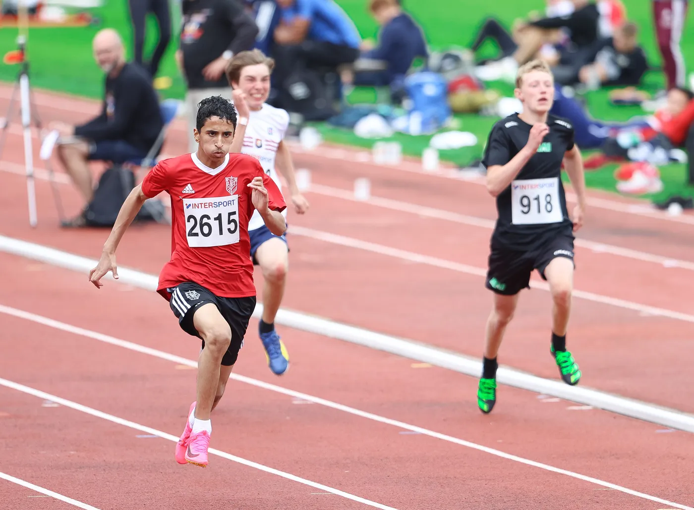 شخص يرتدي قميصًا أحمر مع رقم 2615 يتسابق على المضمار، بينما يطارده عداء آخر يرتدي قميصًا أسود مع رقم 918. في الخلفية، يمكن رؤية مجموعة من المشجعين والرياضيين في بيئة رياضية.