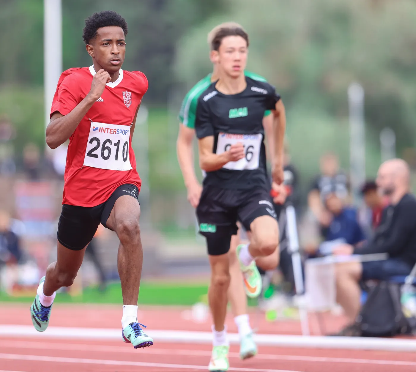 عداء يرتدي قميصًا أحمر يحمل الرقم 2610 في سباق السرعة، بينما يتجاوزه عداء آخر يرتدي قميصًا أسود يحمل الرقم 96. يمكن رؤية بعض الحضور في الخلفية يتابعون المنافسة.