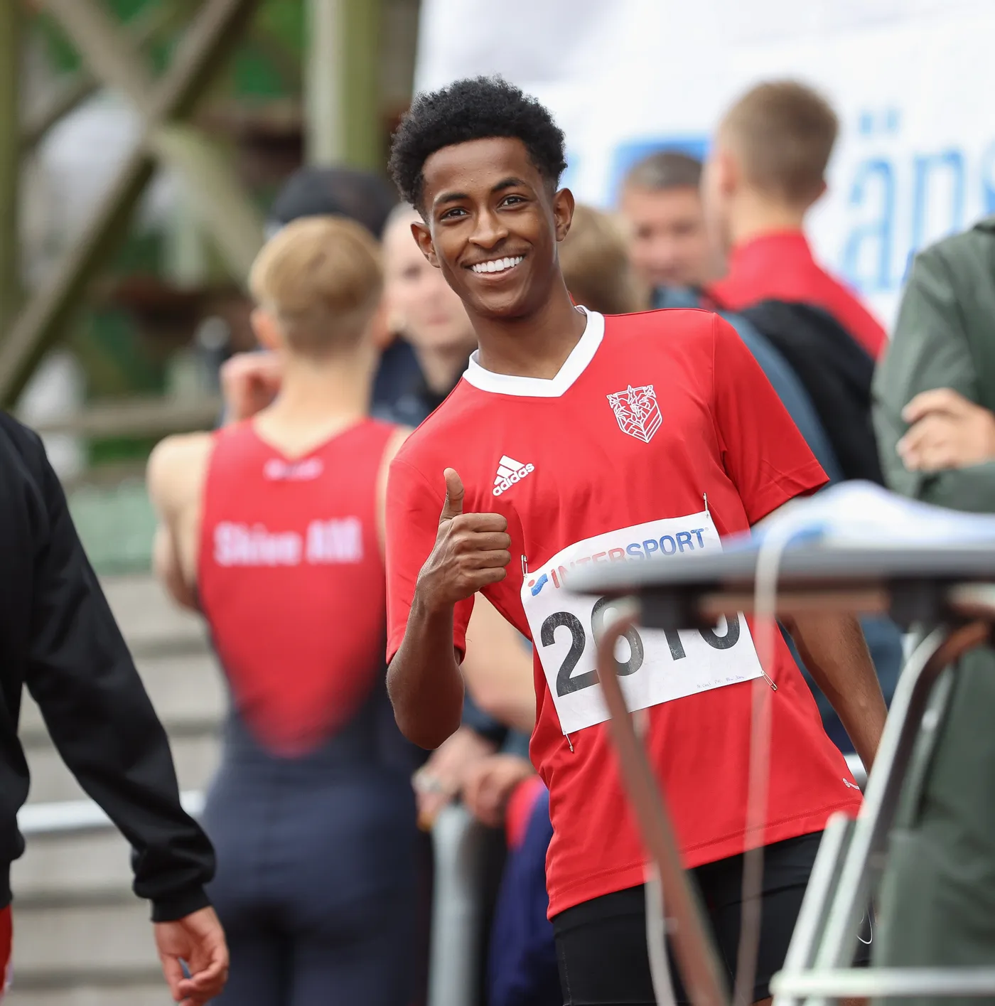 شاب مبتسم يرتدي قميصاً أحمر، يظهر علامة الإعجاب بإبهامه. خلفه مجموعة من الرياضيين والسياق يشير إلى حدث رياضي.