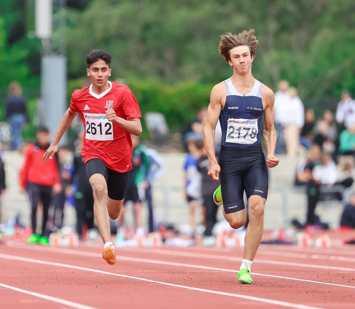 عداء يرتدي قميصًا أحمر يحمل رقم 2612، يتسابق على المضمار، بينما يتفوق عليه عداء آخر يرتدي زيًا رياضيًا أزرق وأسود مع رقم 2178. تظهر في الخلفية مجموعة من المشجعين والرياضيين.