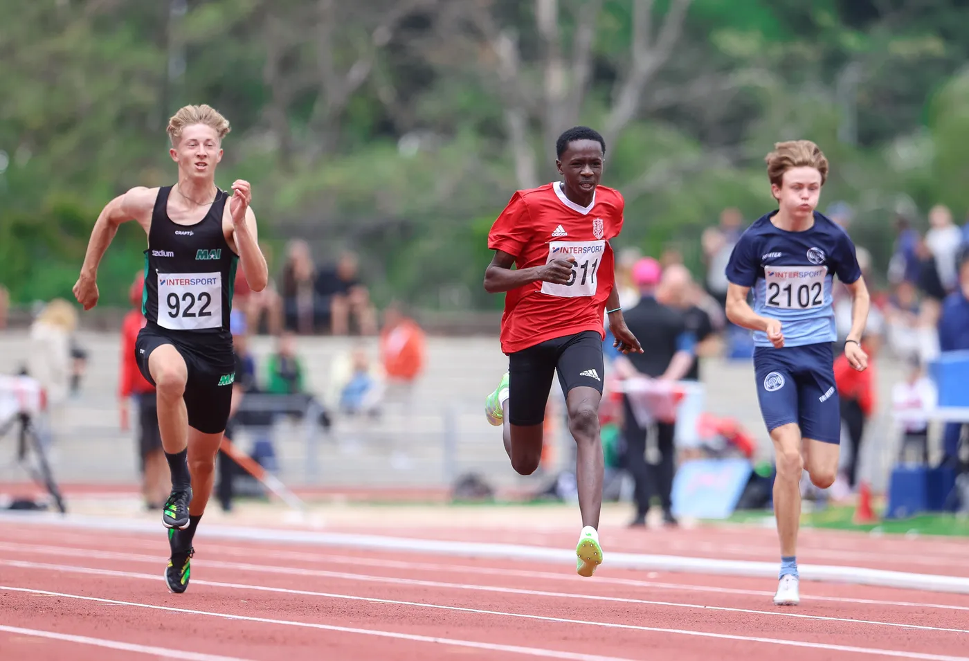 ثلاثة عدّائين يتسابقون على حلبة رياضية. العدّاء في الوسط يرتدي قميصًا أحمر ويبدو أنه يتقدم على الآخرين.