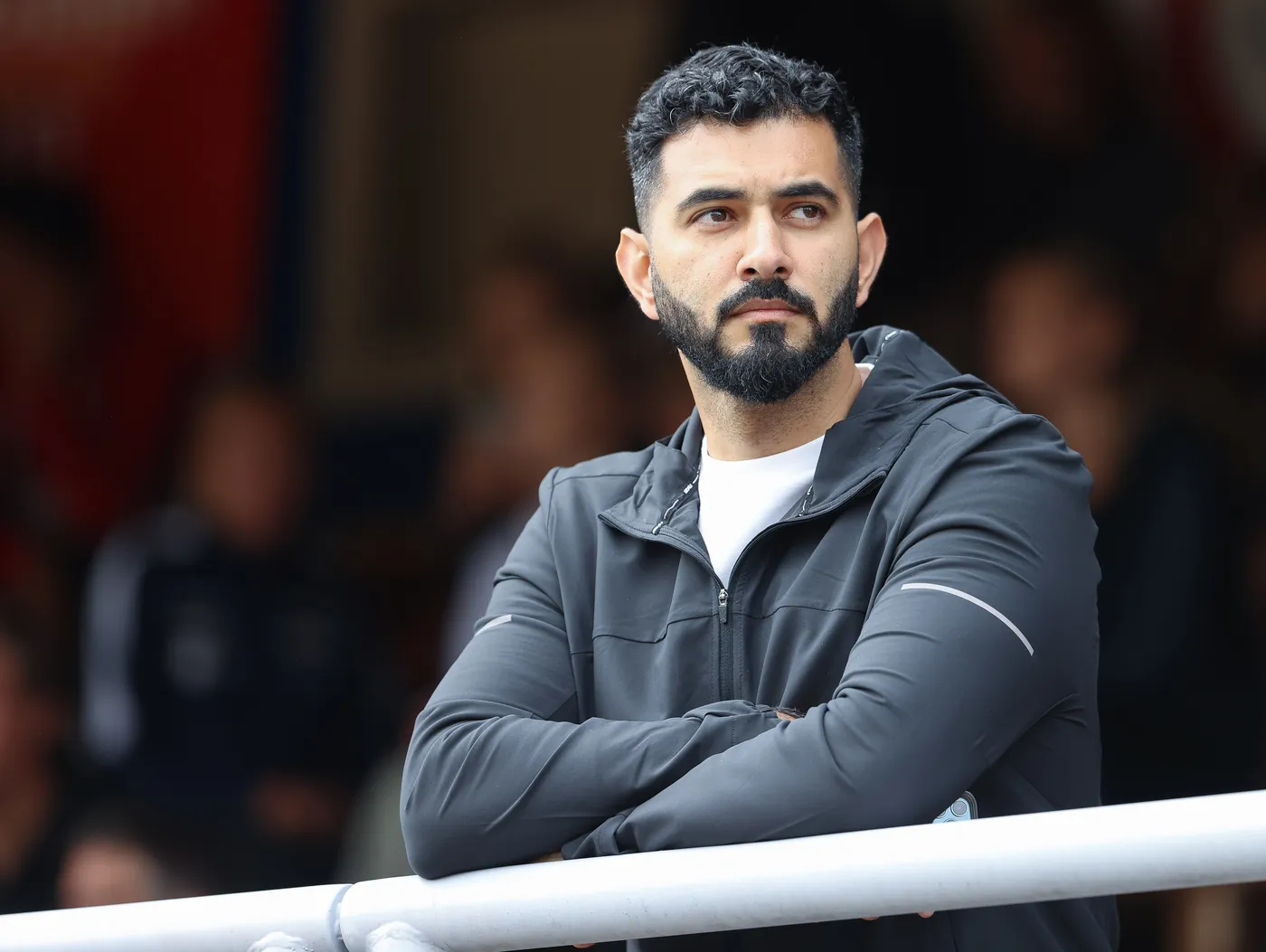 رجل ذو لحية وشعر مجعد يقف بجانب درابزين، مرتديًا سترة رياضية سوداء وقميص أبيض، ويبدو عليه التركيز والاهتمام. خلفه، تظهر ملامح مشجعين في الخلفية.