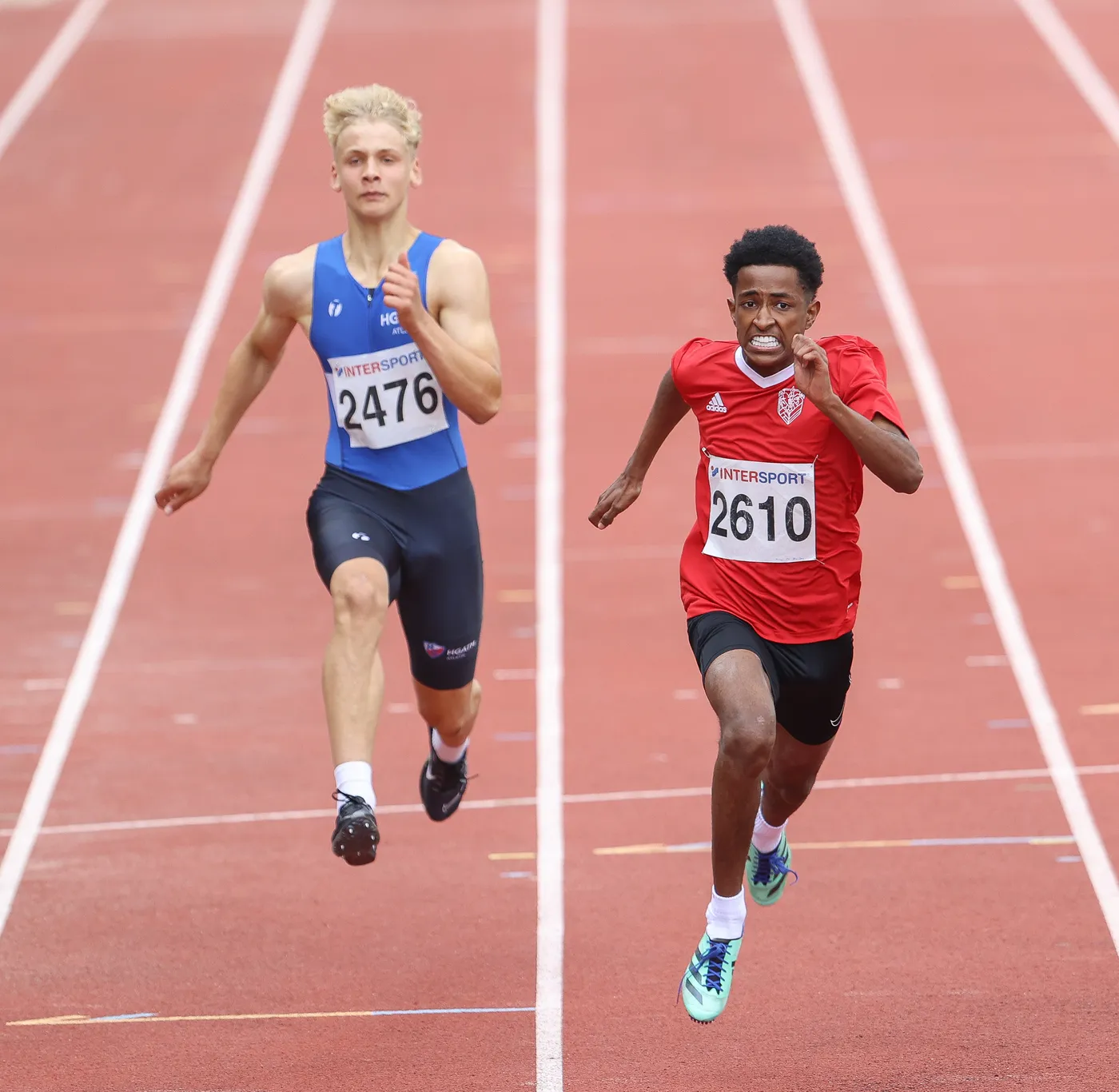 عداءان يتنافسان في سباق على مضمار مخصص، حيث يرتدي الأول تيشرت أزرق برقم 2476، بينما يرتدي الآخر تيشرت أحمر برقم 2610. يبدو أن العداء في التيشرت الأحمر يتقدم في السباق.