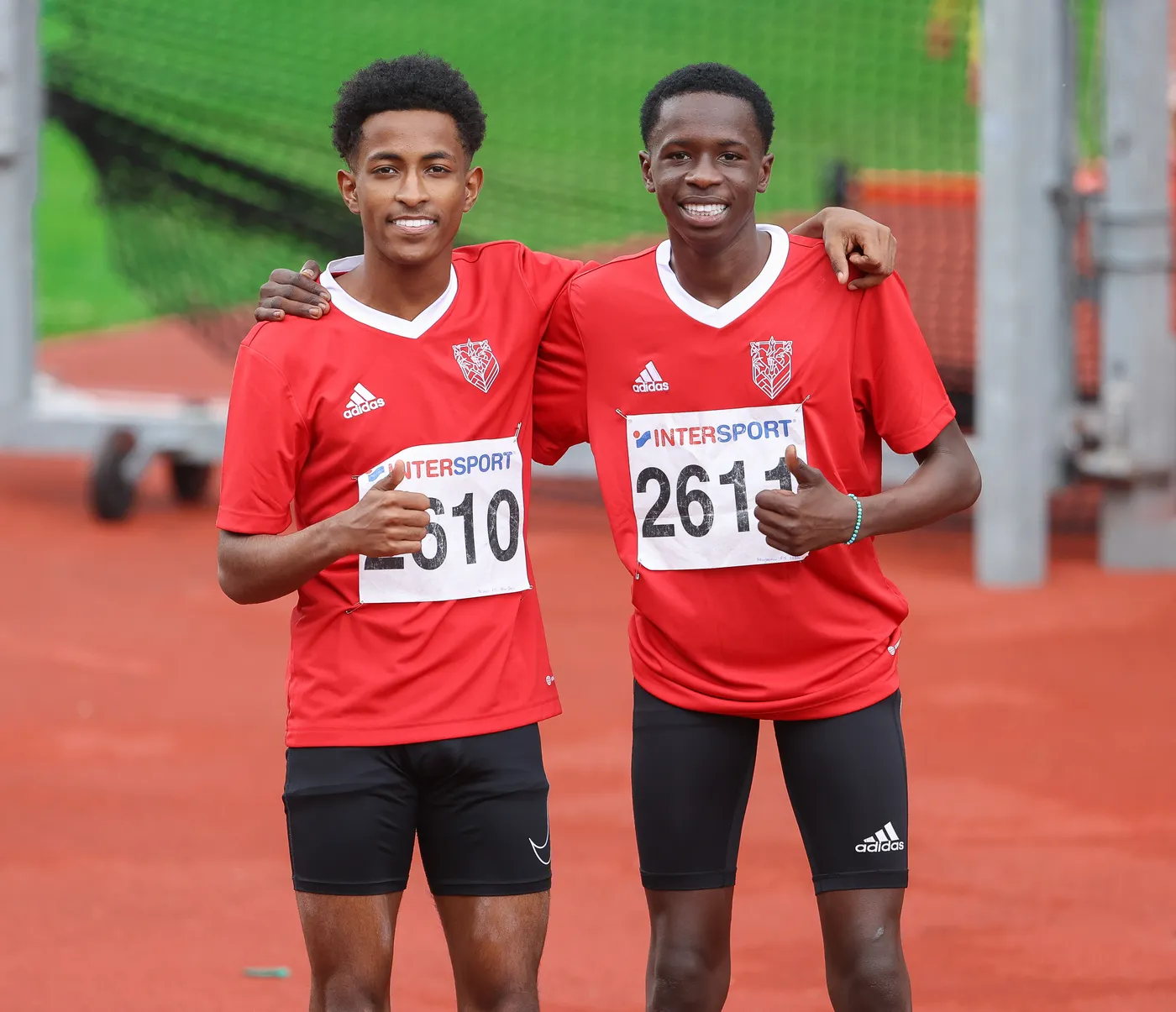 شابان يرتديان قمصانًا حمراء ويبتسمان أثناء وقوفهما على مضمار رياضي. يظهر كل منهما مع رقم على صدره، وكلاهما يحمل إصبعه السبابة بشكل مرفوع في إشارة للنجاح.