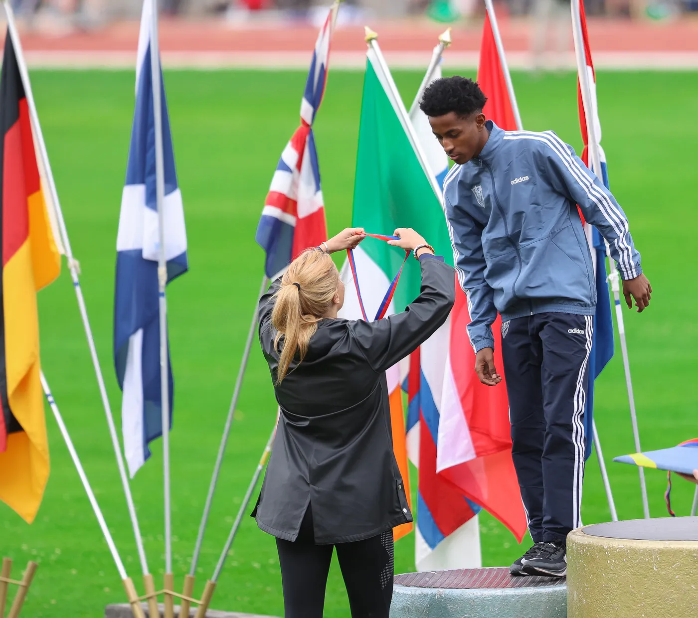شخصان في ساحة رياضية، حيث تقوم امرأة بربط ميدالية حول رقبة شاب واقف على منصة. تتواجد أعلام متعددة خلفهما، مما يعبر عن حدث دولي أو بطولة رياضية.
