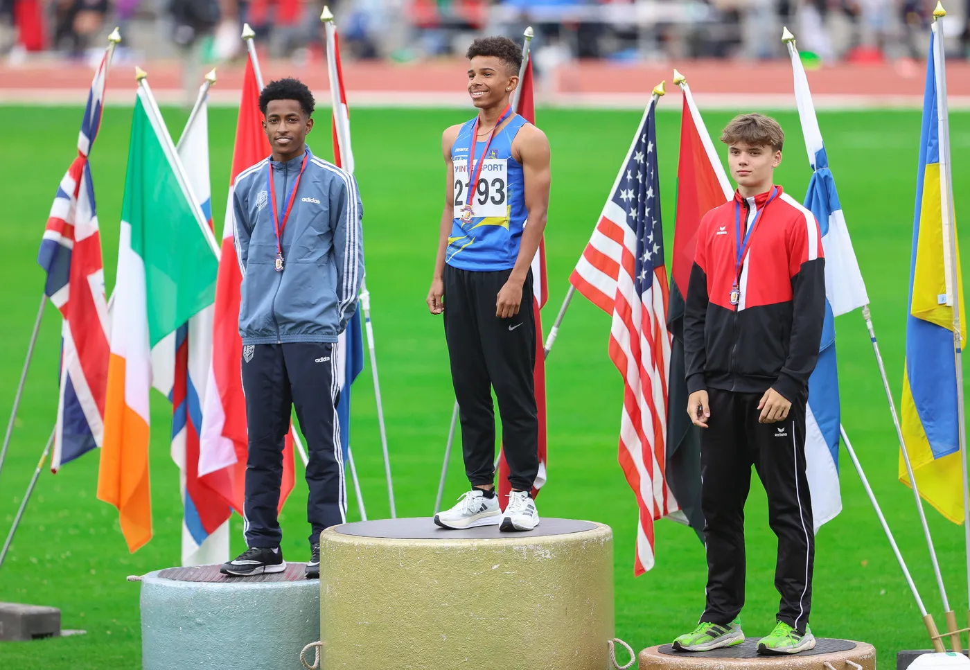 يظهر ثلاثة رياضيين يقفون على منصات التتويج بعد المنافسة، يرتدون ميداليات. الرياضي في المنتصف يبتسم ويرتدي زي رياضي أزرق، بينما الرياضيان على الجانبين يرتديان زيًا باللونين الرمادي والأسود. خلفهم تتواجد عدة أعلام لدول مختلفة.