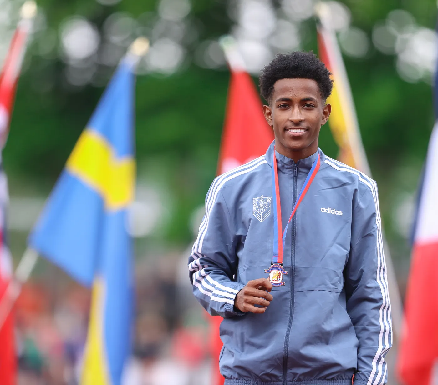 شاب مبتسم يرتدي جاكيت رياضي رمادي يحمل ميدالية حول عنقه. تظهر خلفه أعلام ملونة متعددة تمثل دولًا مختلفة.