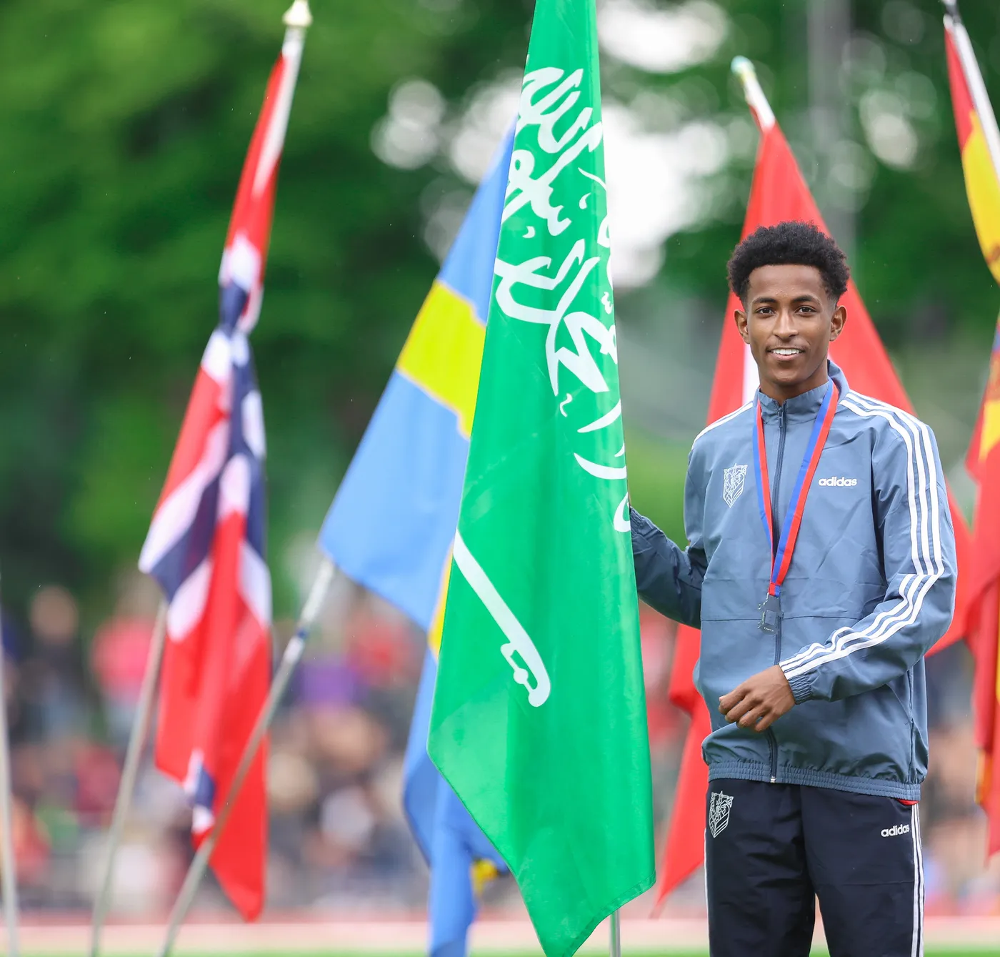شاب يحمل علمًا أخضر مكتوبًا عليه باللغة العربية، بينما تقف أعلام متعددة في الخلفية. يبدو الشاب مبتسمًا ويرتدي ملابس رياضية بجيوب جانبية.
