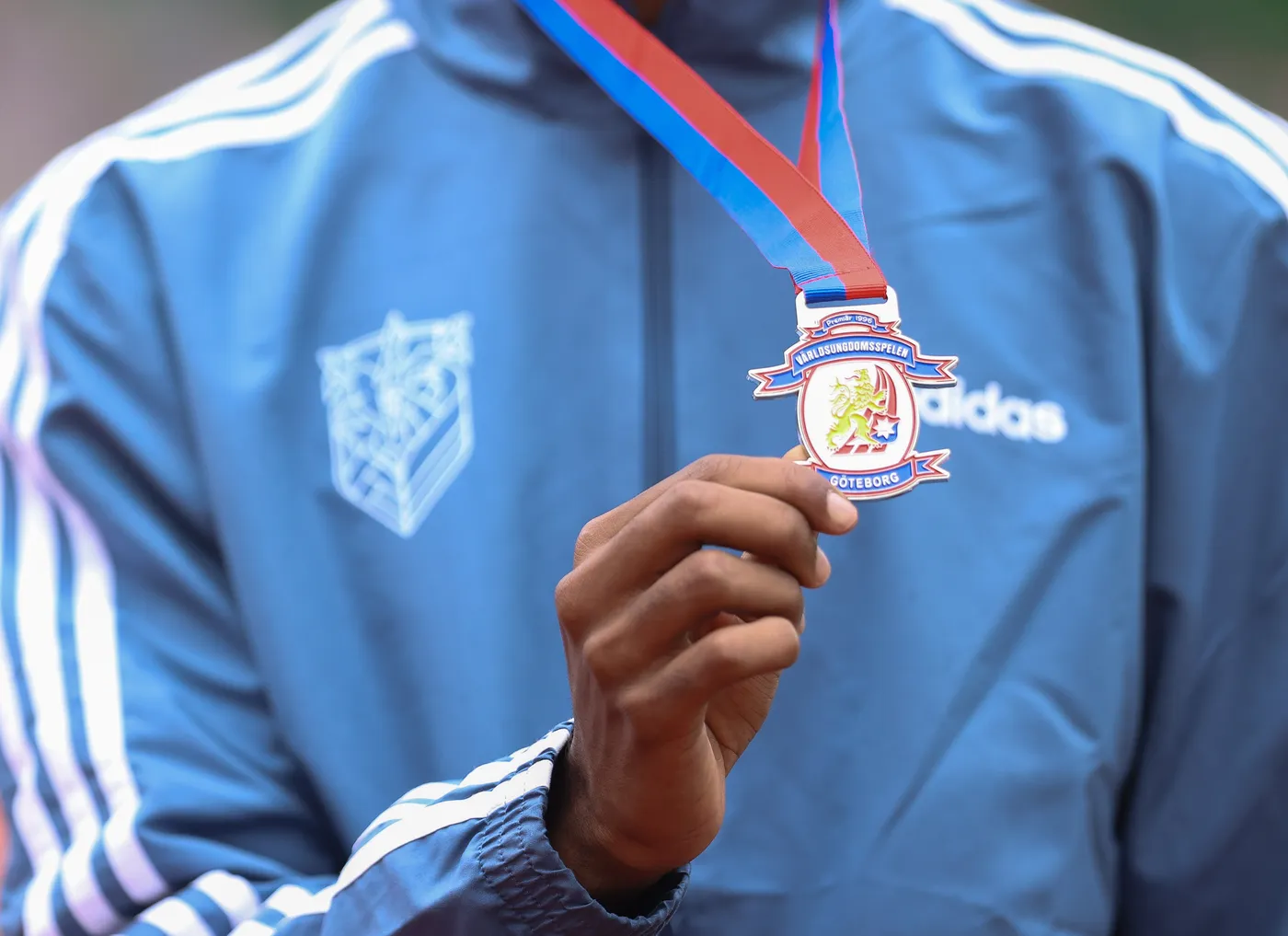 شخص يحمل ميدالية رياضية تتدلى من شريط ذو لونين، الأزرق والأحمر، ويرتدي سترة رياضية زرقاء مع خطوط بيضاء. الميدالية تحمل شعار وقد كتب عليها 
