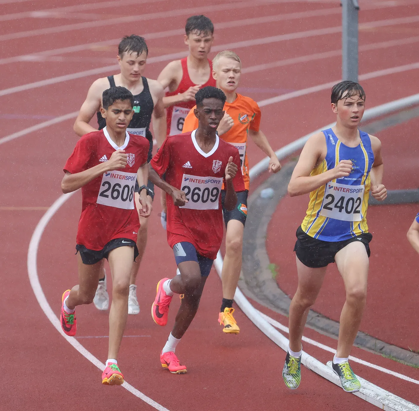 مجموعة من عدائي المراهقين يتسابقون على حلبة تحت المطر، يرتدي بعضهم قمصان حمراء وأرقام على صدورهم، بينما يرتدي الآخرون ملابس بألوان مختلفة. التعبيرات على وجوههم تعكس التركيز والجهد أثناء المنافسة.