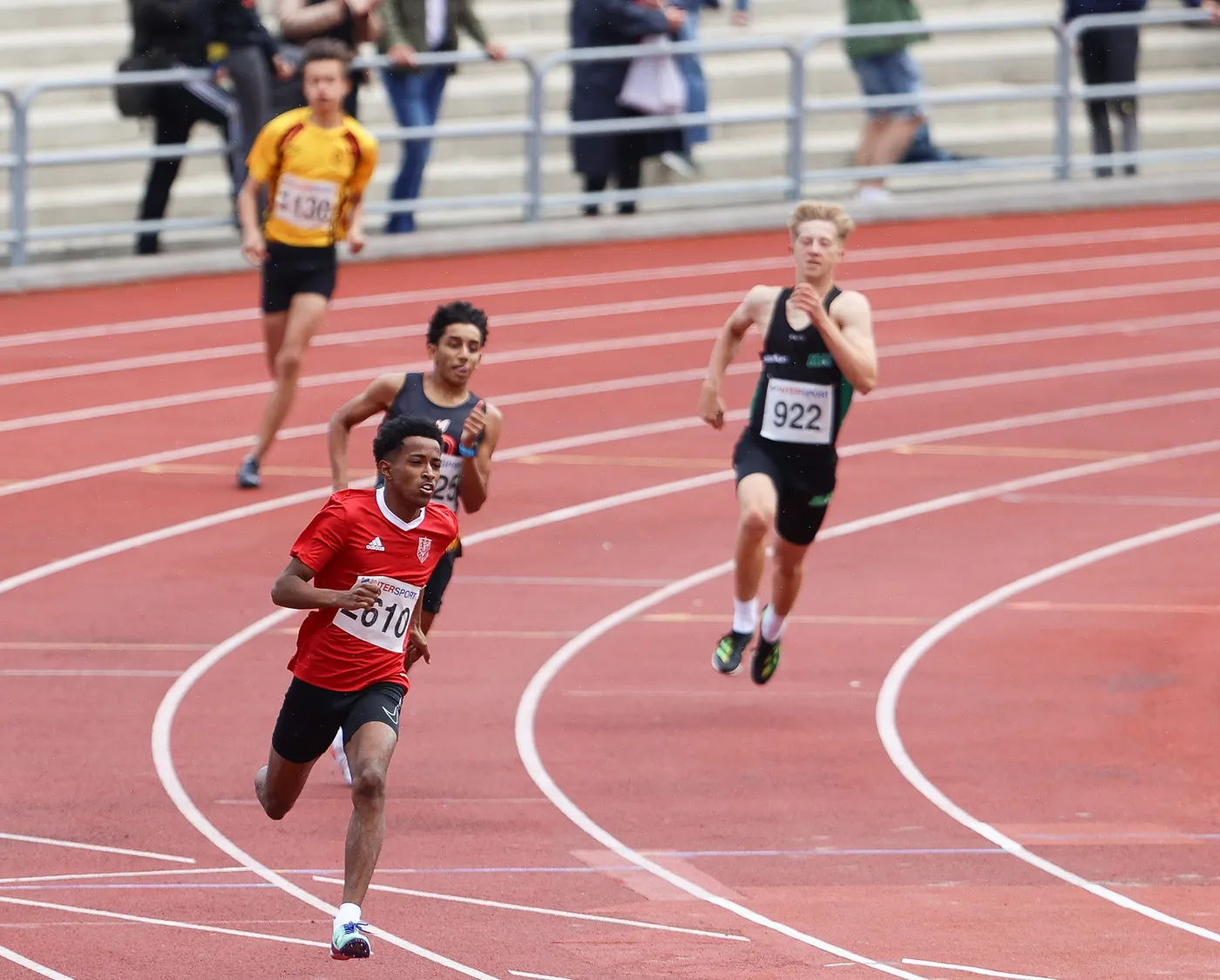 عداء يرتدي قميصًا أحمر يحمل الرقم 610 يجري بسرعة على حلبة سباق بينما يتفوق على عدائين آخرين. هناك متسابقون آخرون في الخلف، والسباق يبدو شديد التنافس.