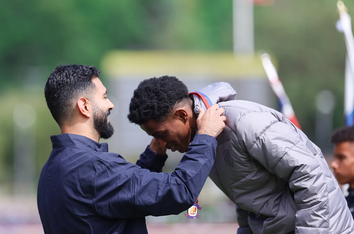 مدرب يكافئ لاعبًا بفوز بعد وضع ميدالية على عنقه بينما يبتسم اللاعب، مع وجود لاعبين آخرين في الخلف. الأجواء تحمل شعورًا بالاحتفال والإنجاز.