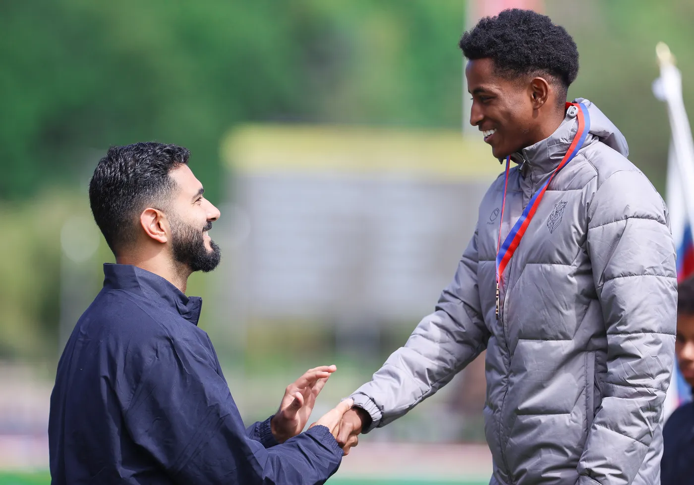 رجل ذو لحية وابتسامة يرتدي سترة داكنة shakes hands with another young man wearing a gray padded jacket adorned with a medal around his neck, both smiling at each other in a sunny outdoor setting.