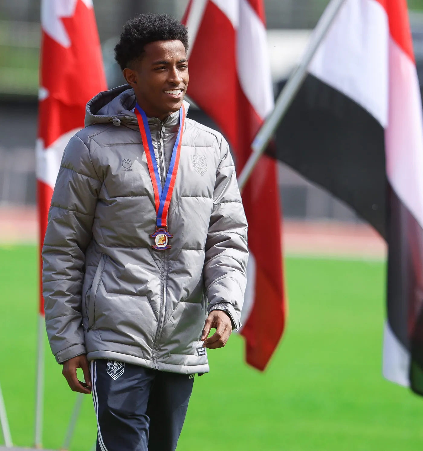 شاب يرتدي سترة رمادية ويحمل ميدالية على صدره، مبتسم وهو يسير على أرضية خضراء محاطًا بأعلام ملونة.