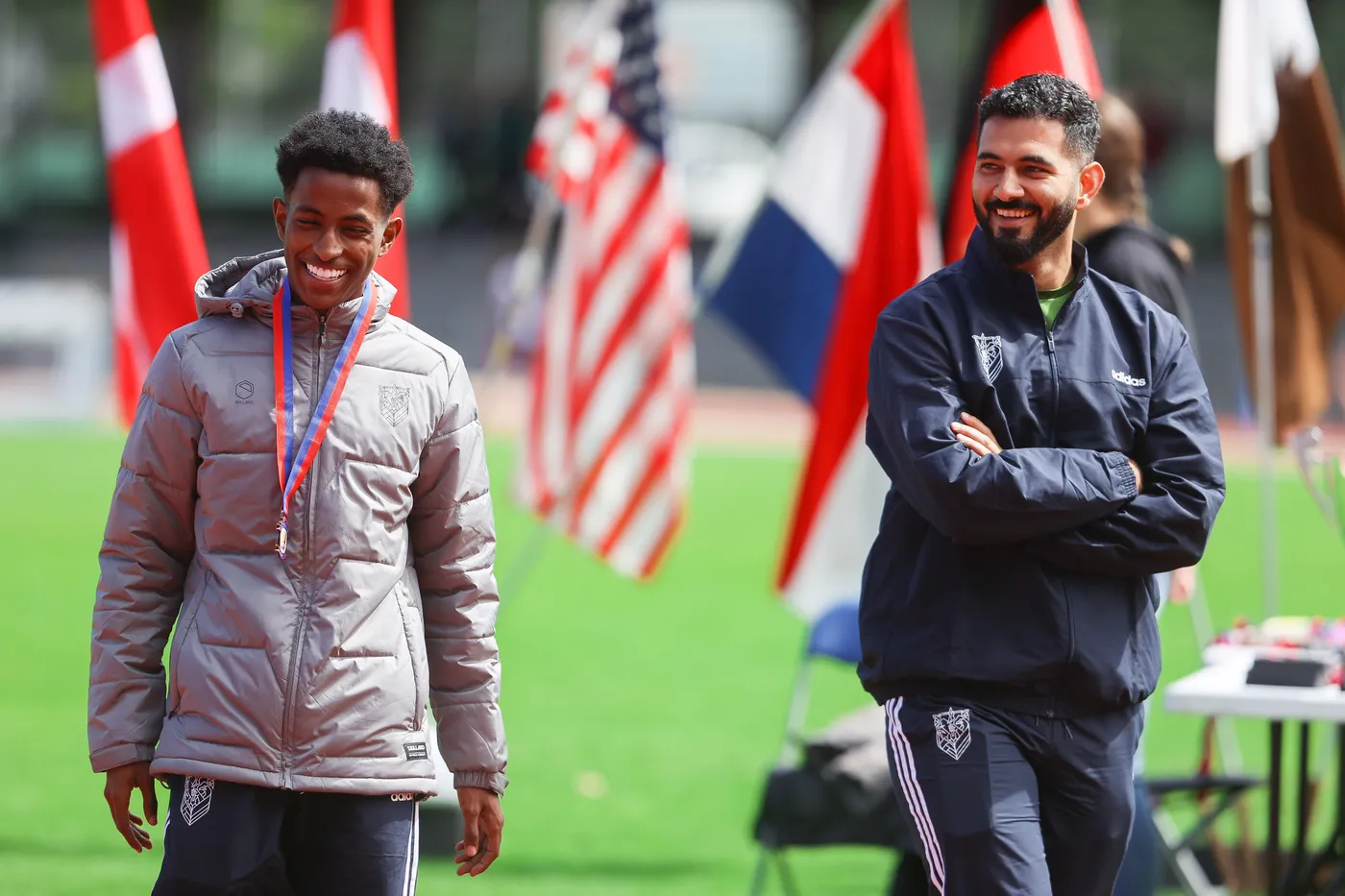 شاب مبتسم يرتدي سترة رمادية ويحمل ميدالية على رقبته، يقف بجوار رجل ذو لحية قصيرة يرتدي سترة داكنة. خلفهم، تتواجد أعلام متعددة تتمايل في الهواء.