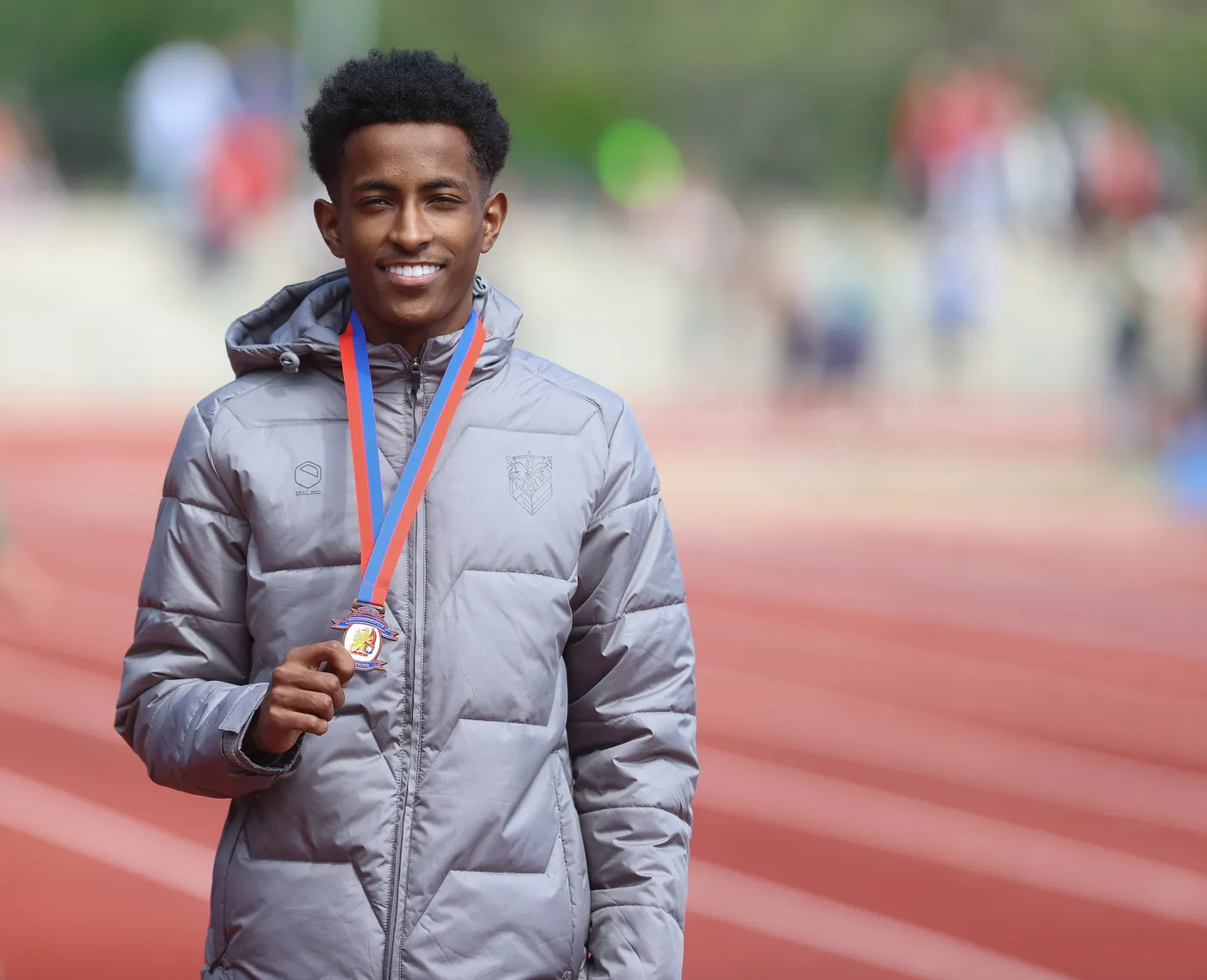 شاب مبتسم يرتدي سترة رمادية ويحتفظ بميدالية رياضية تتدلى حول عنقه، يقف على مضمار رياضي. الخلفية تظهر مجموعة من الأشخاص في أجواء رياضية.