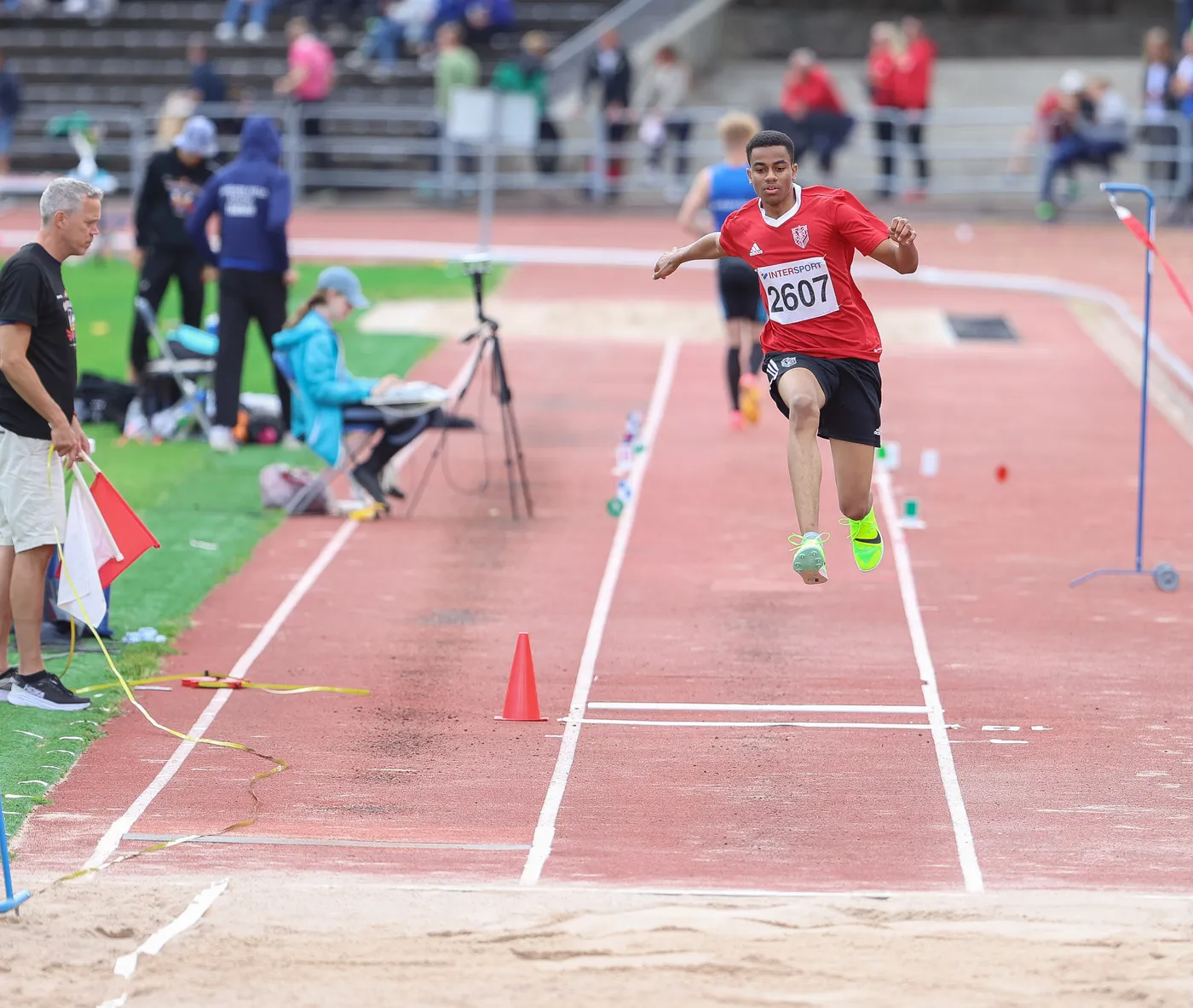 شاب يرتدي قميصاً أحمر ورقم 2607، يقفز في مسابقة الوثب الطويل. يظهر على حلبة رياضية مع جمهور في الخلفية وأشخاص يلاحظون المنافسة.