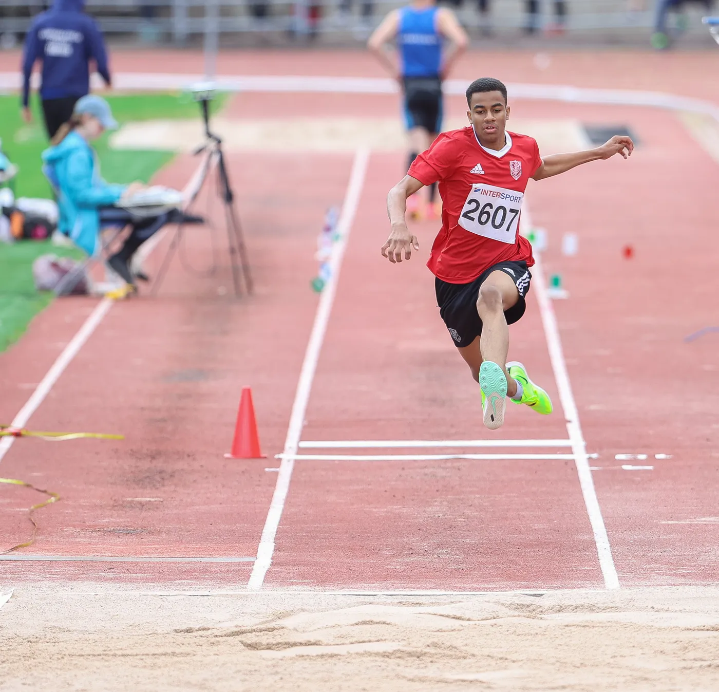 عداء يرتدي قميصًا أحمر وبه رقم 2607، يقفز في الهواء فوق خط الرمز الخاص بقفز المسافة في مضمار رياضي. في الخلفية، يُمكن رؤية بعض المتفرجين ومعدات التسجيل.