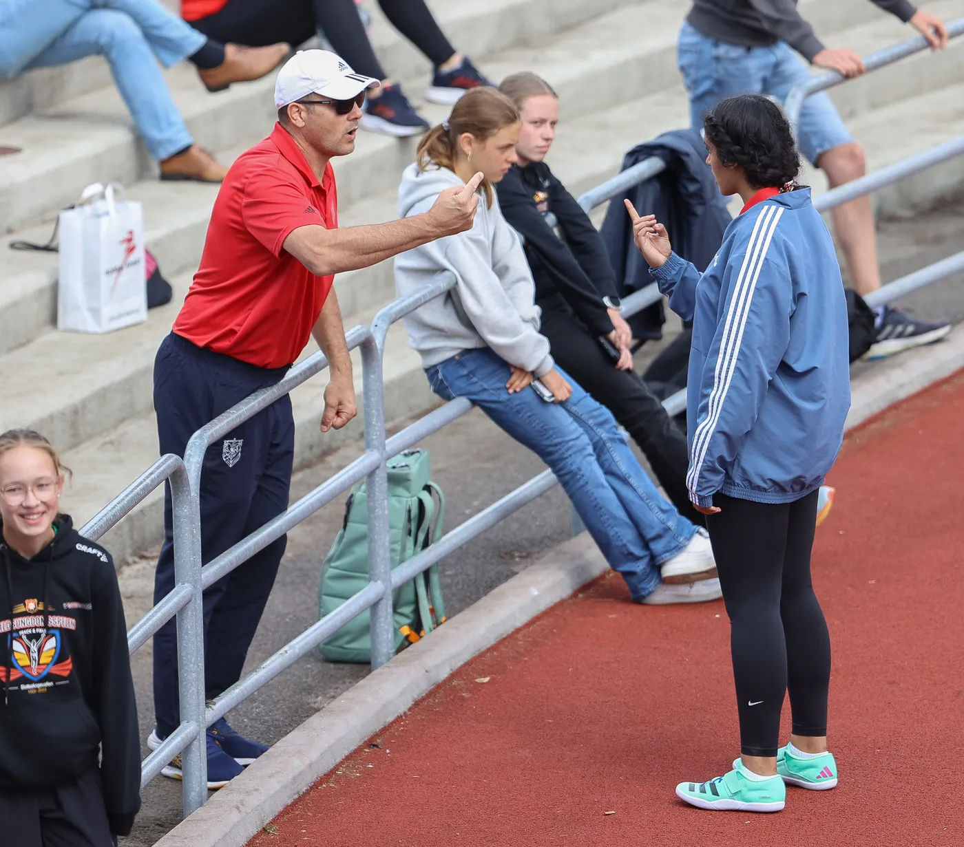 رجل يرتدي قميصًا أحمر ويقف بجانب سياج يتحدث بحماس مع شابة ترتدي سترة زرقاء ومقفولة يدها، بينما يجلس مجموعة من الفتيات في الخلف. الجو يبدو غير رسمي، مع تركيز واضح بين الطرفين.