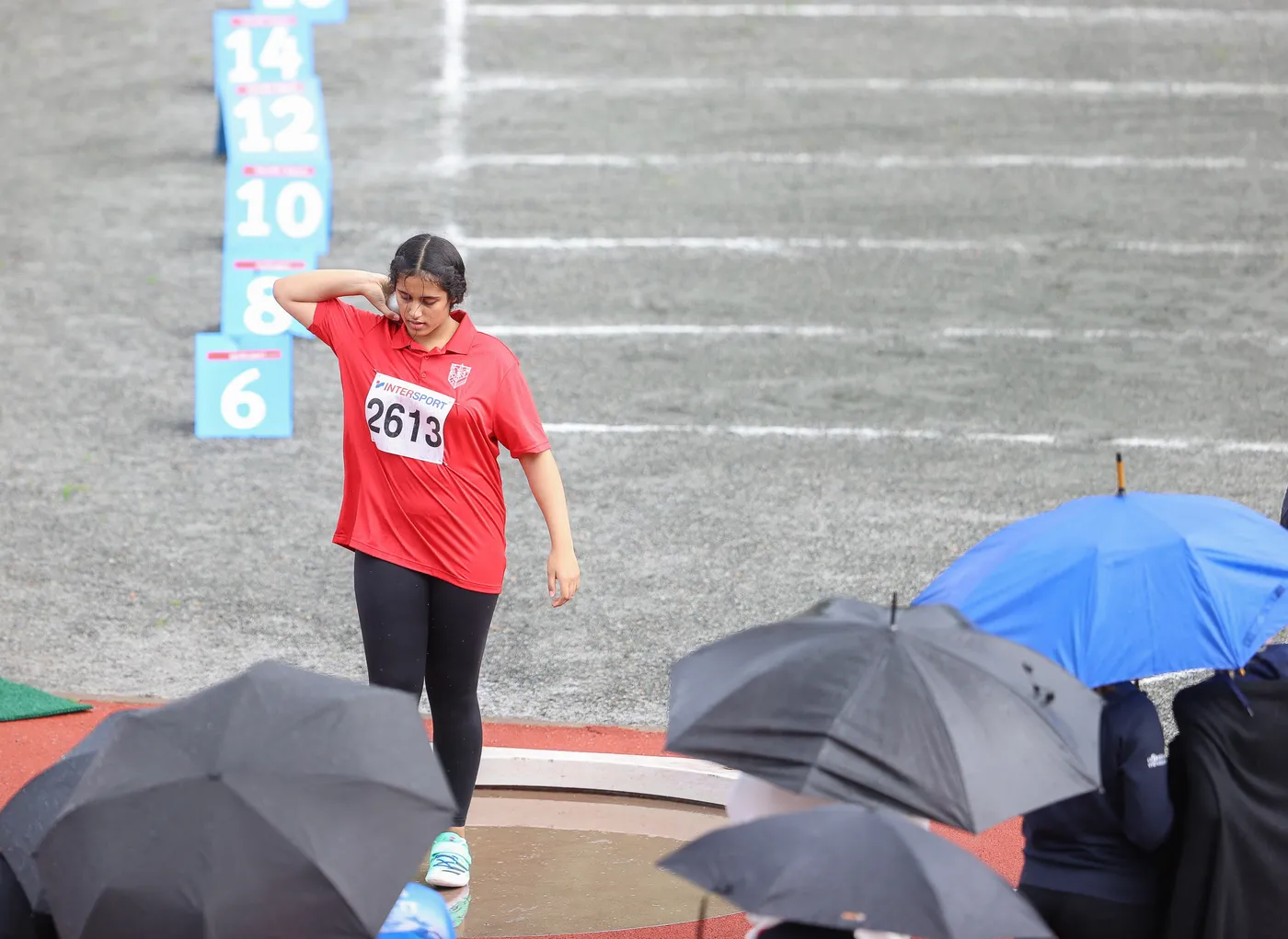 امرأة ترتدي قميصًا أحمر يحمل رقم 2613، تتجه نحو منطقة المنافسة في ملعب مبلّل بالمطر. الخلفية تحتوي على علامات ملونة على الأرض تحت المطر، مع مظلات سوداء وزرقاء تحمي المتفرجين من الأمطار.