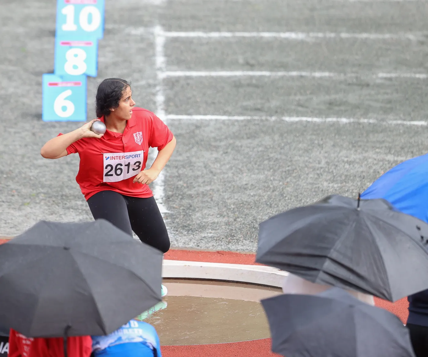 شابة ترتدي قميصاً أحمر وتستعد لرمي كرة معدنية في مضمار رياضي. يظهر في الخلفية علامات الأرقام مع الأمطار في المكان.