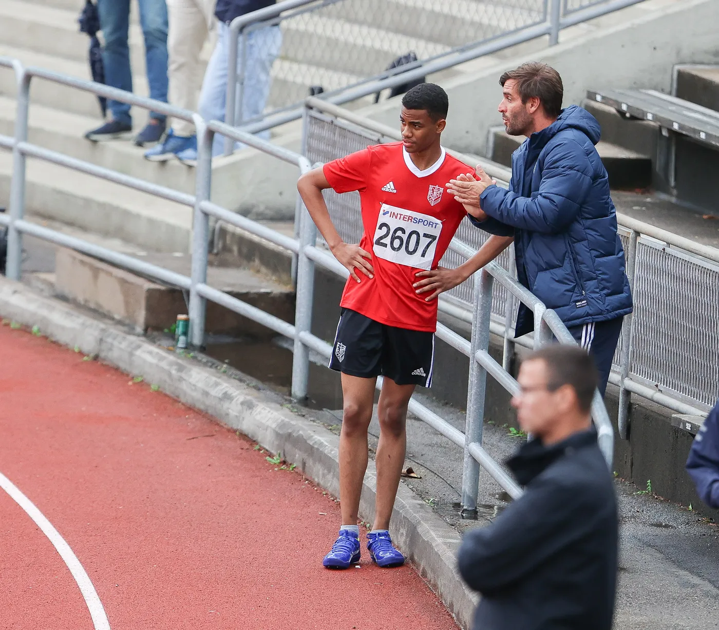 شاب يرتدي قميصًا رياضيًا أحمر يحمل الرقم 2607، يقف بجانب مدربه الذي يبدو أنه يعطيه توجيهات. الخلفية تحتوي على بعض المتفرجين، فيما يوجد مضمار رياضي يتضح من لونه الأحمر.