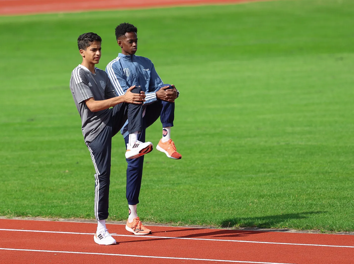 شخصان يمارسان تمارين الإحماء على المضمار. الواحد منهما يرفع ساقه تجاه صدره بينما الآخر يساعده في التوازن. الأرضية مغطاة بالعشب الأخضر والمضمار محاط بأرضية حمراء.