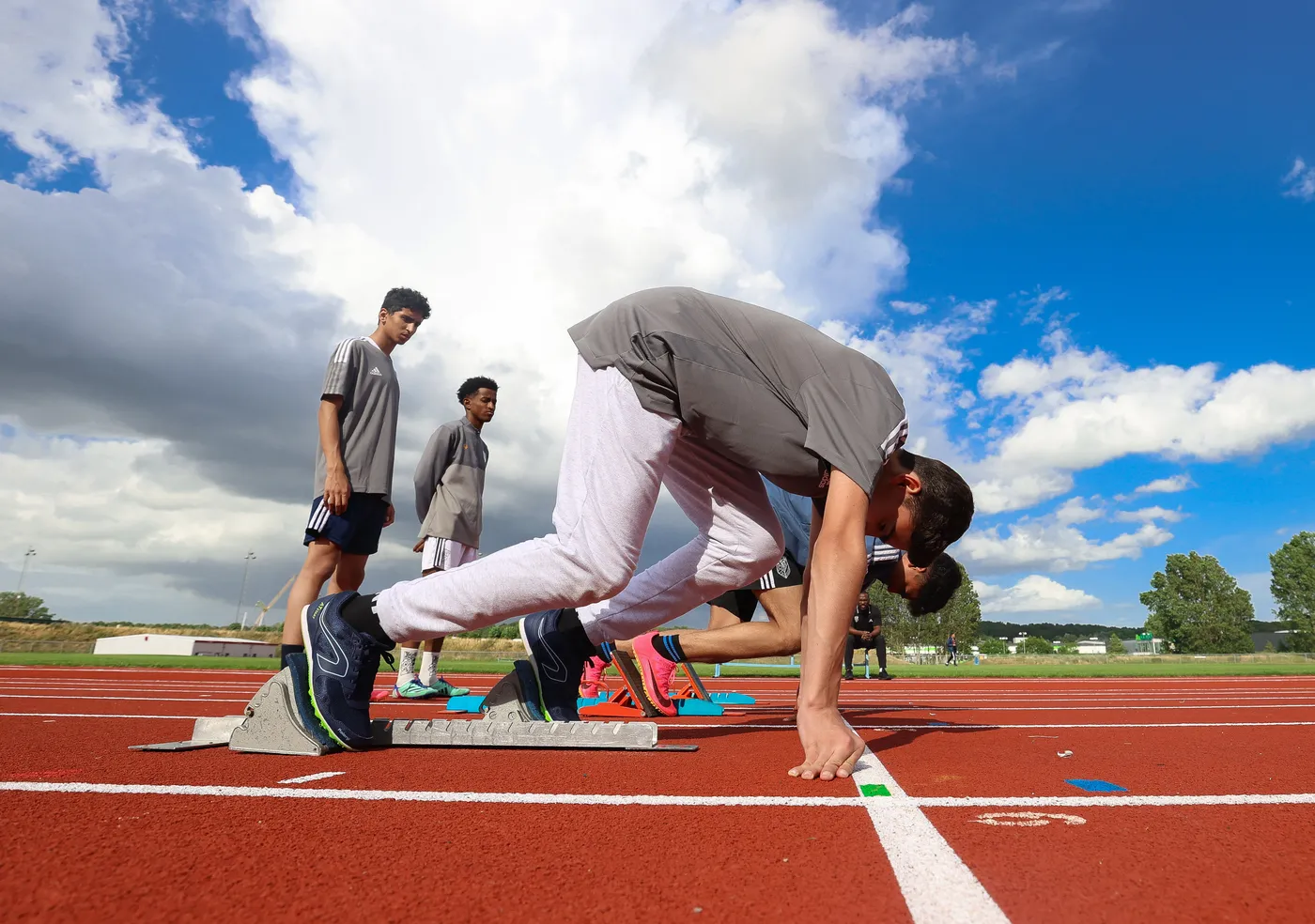مجموعة من الشباب يرتدون زي رياضي يقفون على مسار الجري. شخص واحد في الوضع المنخفض لبدء السباق، بينما ينظر الآخرون بتعابير التركيز. السماء زرقاء وتحمل بعض السحب.