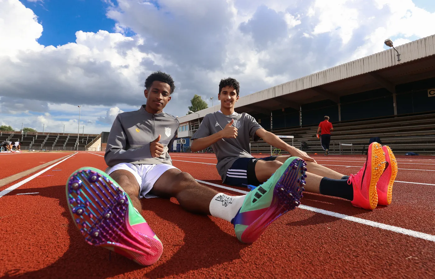 شابان يجلسان على بودرة حمراء لملعب رياضي، يرتديان تيشيرتات رياضية، مع تعابير مبتهجة، يرفعان إبهاميهما إلى الأعلى. في الخلفية، يظهر ملعب مع مقاعد.