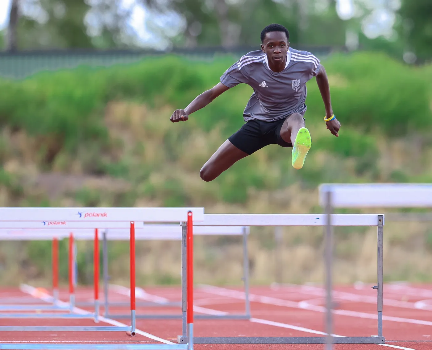 عداء يتخطى حواجز أثناء المنافسة في مضمار رياضي. يرتدي قميصًا رماديًا وشورتًا أسود، ويظهر في قفزة قوية بينما تتواجد الحواجز أمامه.