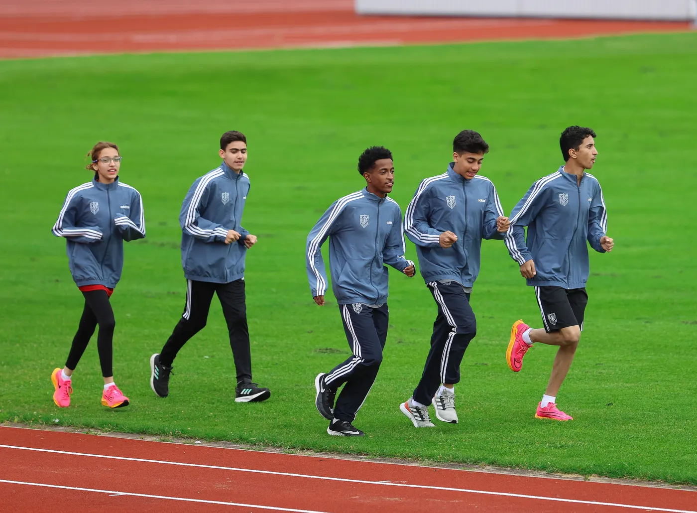 مجموعة من الشباب والأطفال يرتدون ملابس رياضية رمادية، يجري خمسة منهم على مضمار رياضي محاط بالعشب الأخضر. يعبر بعضهم الابتسامة بينما يتجهون في خط مستقيم.