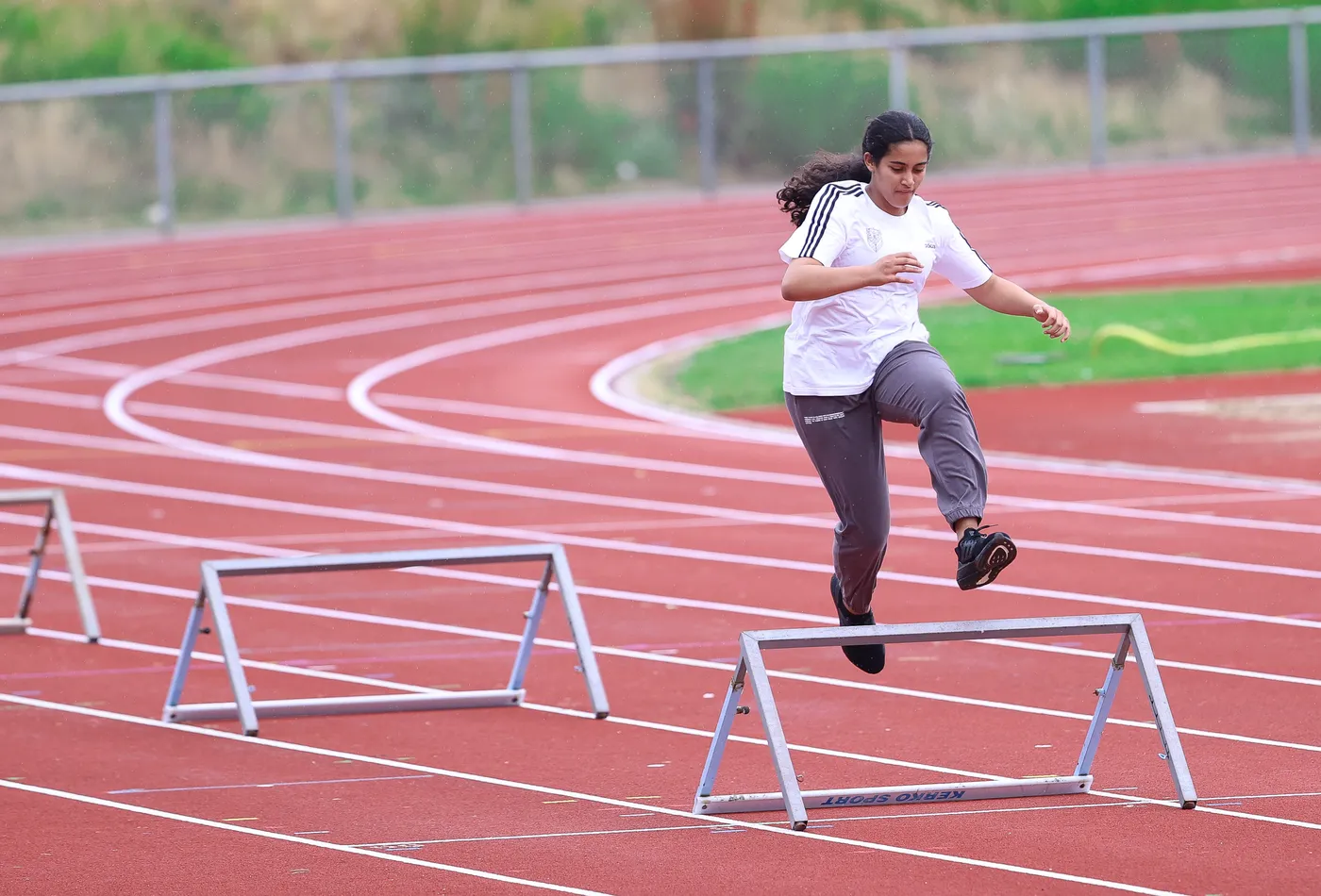 شابة رياضية تتخطى حاجزًا في حلبة مضمار رياضي. ترتدي ملابس رياضية وتظهر عازمة ومركزة في أدائها.