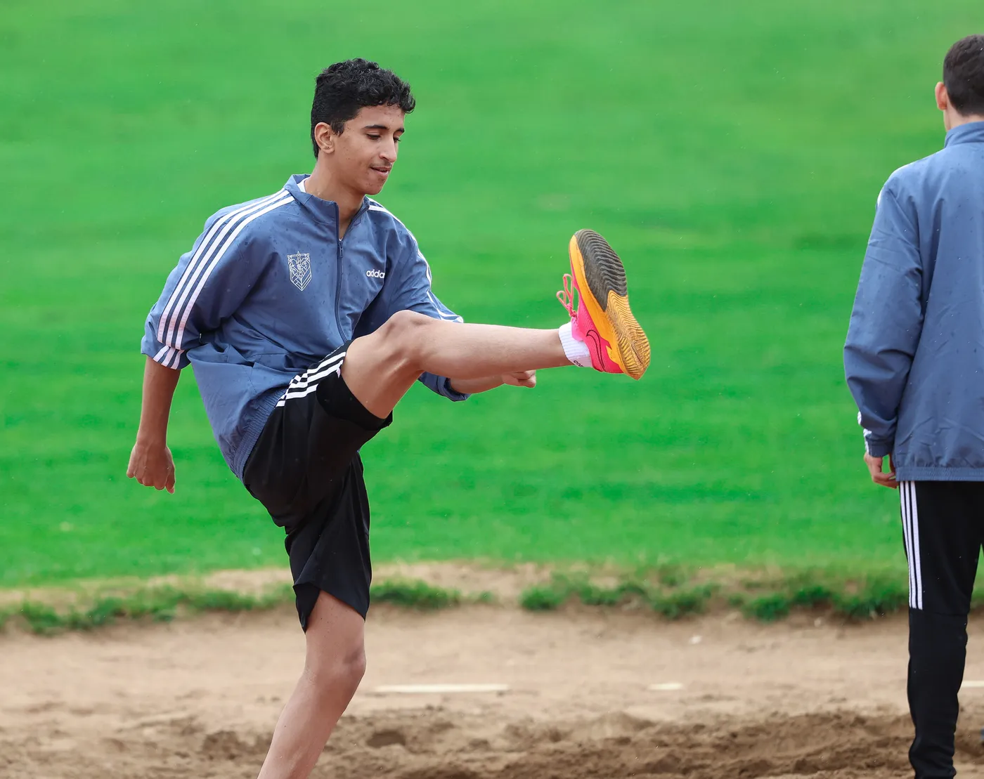 شاب يرتدي ملابس رياضية، يقوم بتمارين الإحماء برفع ساقه. في الخلفية، يوجد شخص آخر يرتدي ملابس رياضية، وكل ذلك يتواجد في ساحة خضراء.