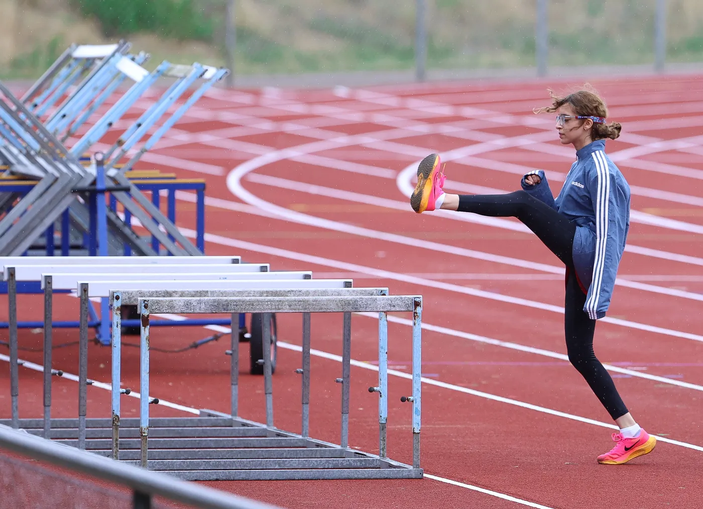 تتدرب فتاة في حلبة رياضية على تخطي الحواجز، حيث ترفع إحدى ساقيها في الهواء بينما تسير على حلبة حمراء. الجو يبدو غائمًا، مما يشير إلى احتمال هطول المطر.