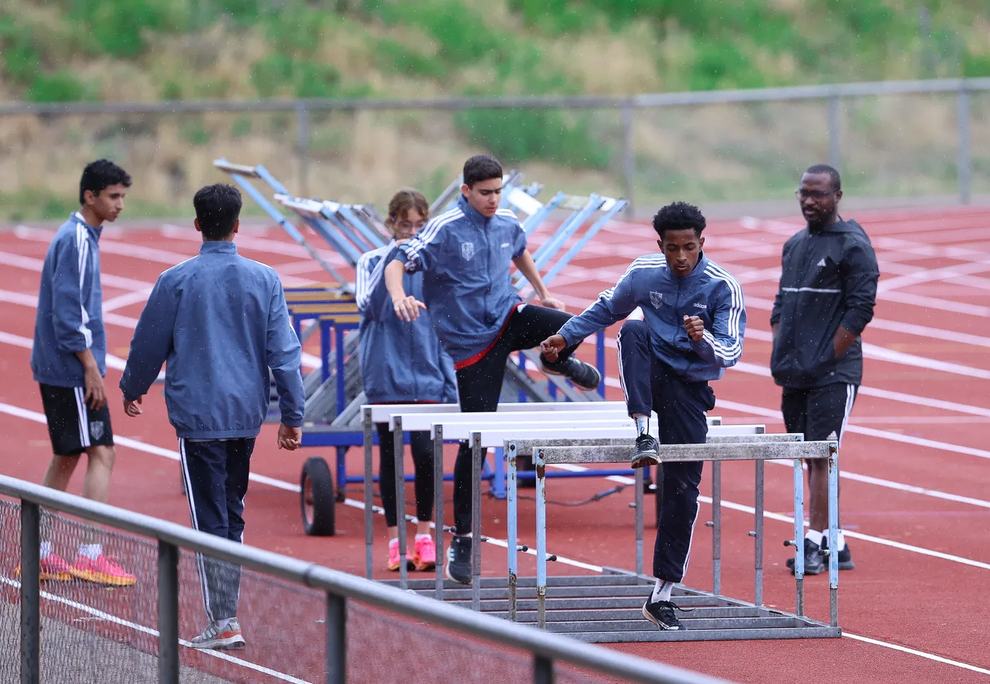 مجموعة من الشباب يرتدون ملابس رياضية يتدربون على سباق الحواجز على حلبة في الهواء الطلق. يظهر بعضهم وهم يتخطون الحواجز بينما يراقبهم آخرون.