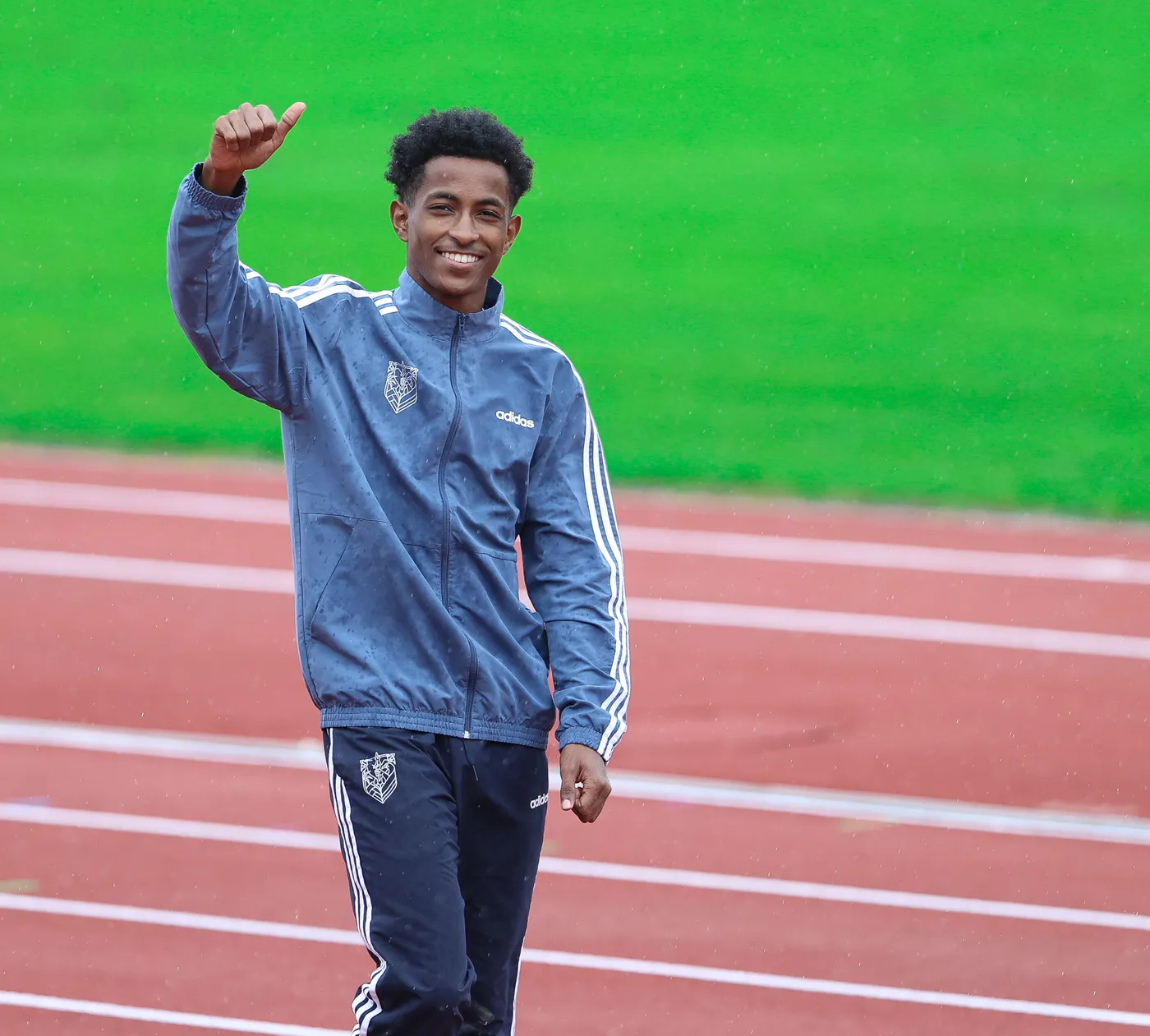 شخص شاب مبتسم يرتدي جاكيت رياضي وسروال رياضي، يسير على مضمار رياضي ويقوم بإشارة الإيجابية بيده. الخلفية مليئة بالعشب الأخضر ومضمار الجري أحمر.