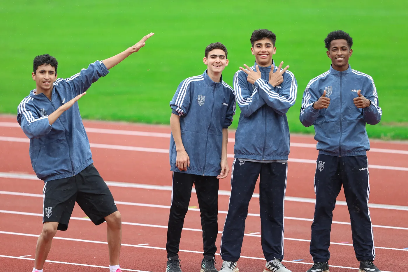 أربعة شباب يرتدون زي رياضي أزرق، يقفون على مضمار رياضي، يبتسمون ويتبادلون الإشارات بإيماءات مرحة. الخلفية تتكون من عشب أخضر ومضمار رياضي.