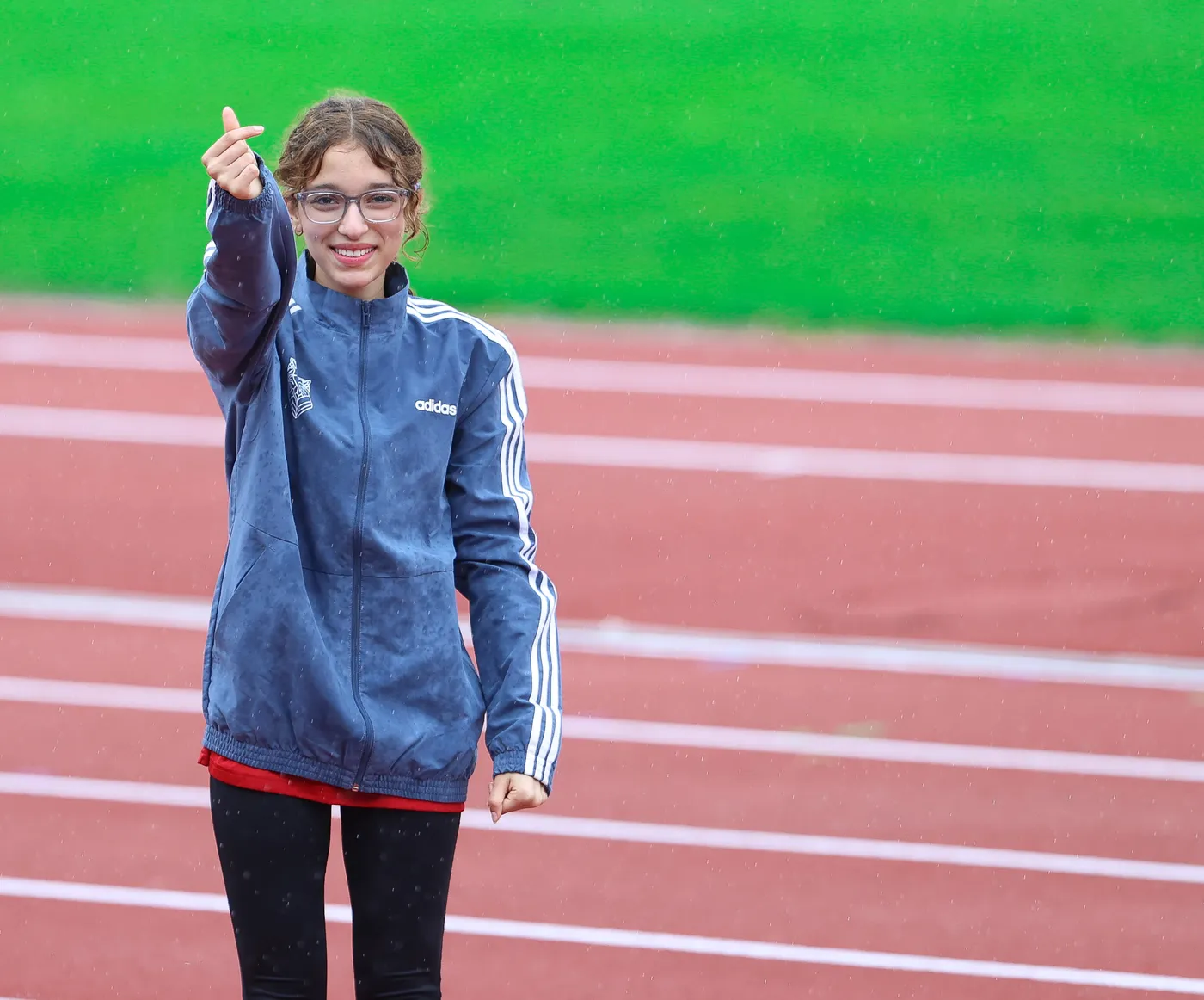 فتاة ترتدي معطف رياضي داكن مع خطوط بيضاء، تبتسم وتقدم إشارة بيدها. خلفها، مضمار رياضي أخضر مع خطوط حمراء تحت المطر.