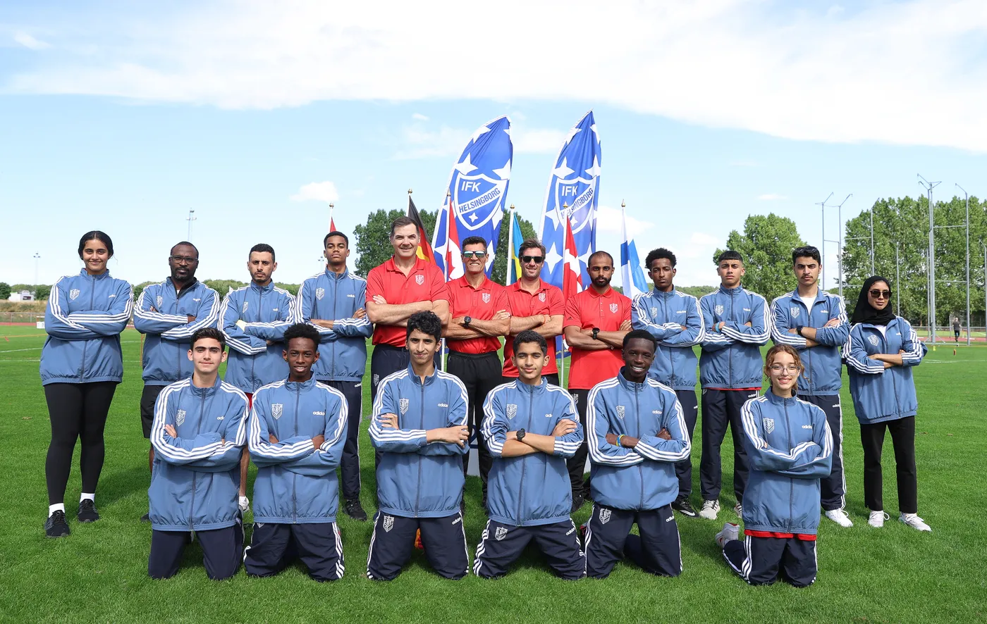 مجموعة من الأشخاص يقفون مبتسمين في ملعب أخضر، يرتدون ملابس رياضية زرقاء، مع بعضهم في الصفوف الأمامية، بينما يقف المدربون في الخلف. هناك أعلام ملونة تشير إلى فريق أو احتفال رياضي في الخلفية.