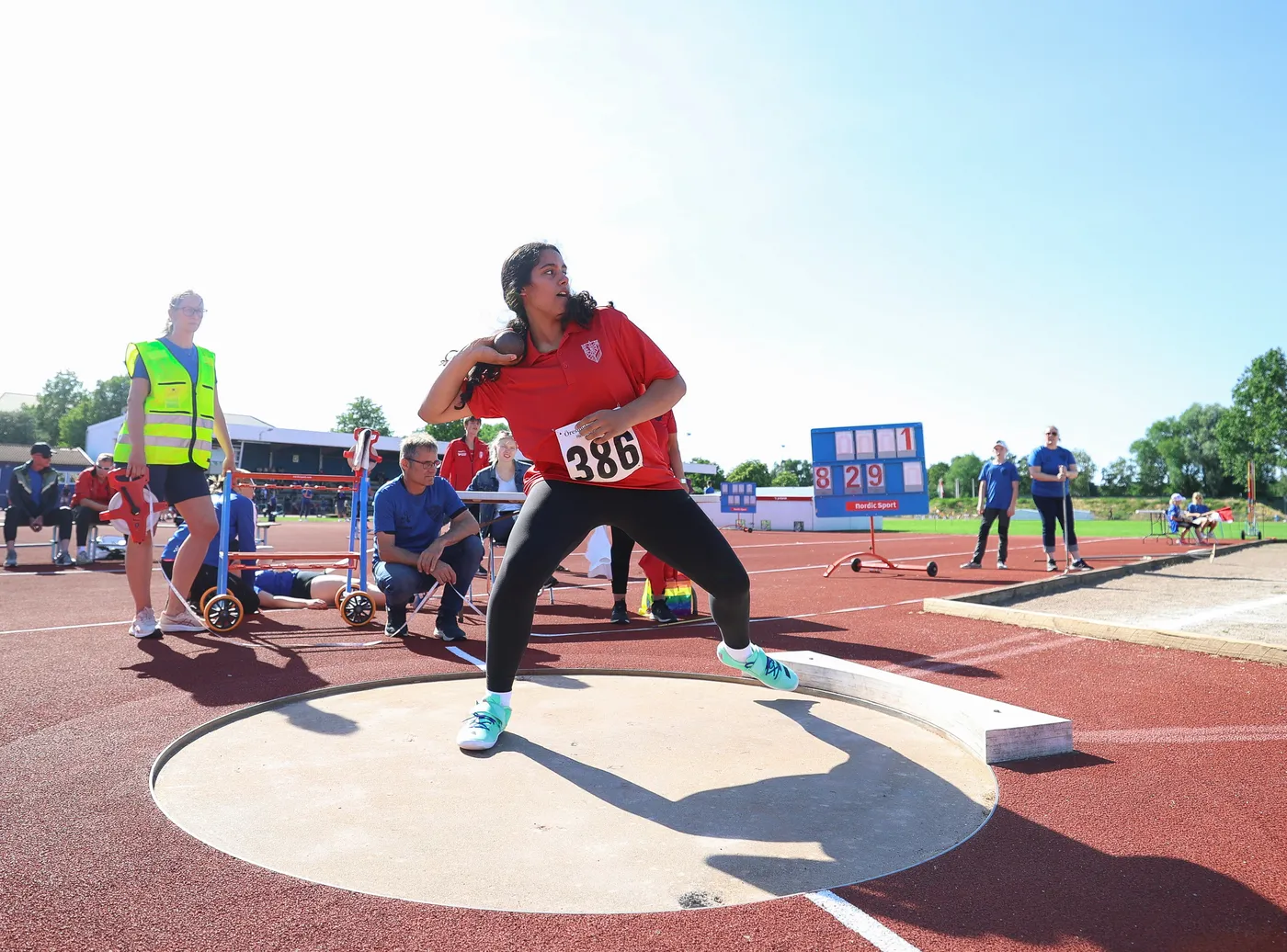 Athlete in a red shirt prepares to throw a shot put at a sporting event. A scoreboard displays scores in the background while spectators and officials watch from the sidelines under clear blue skies.