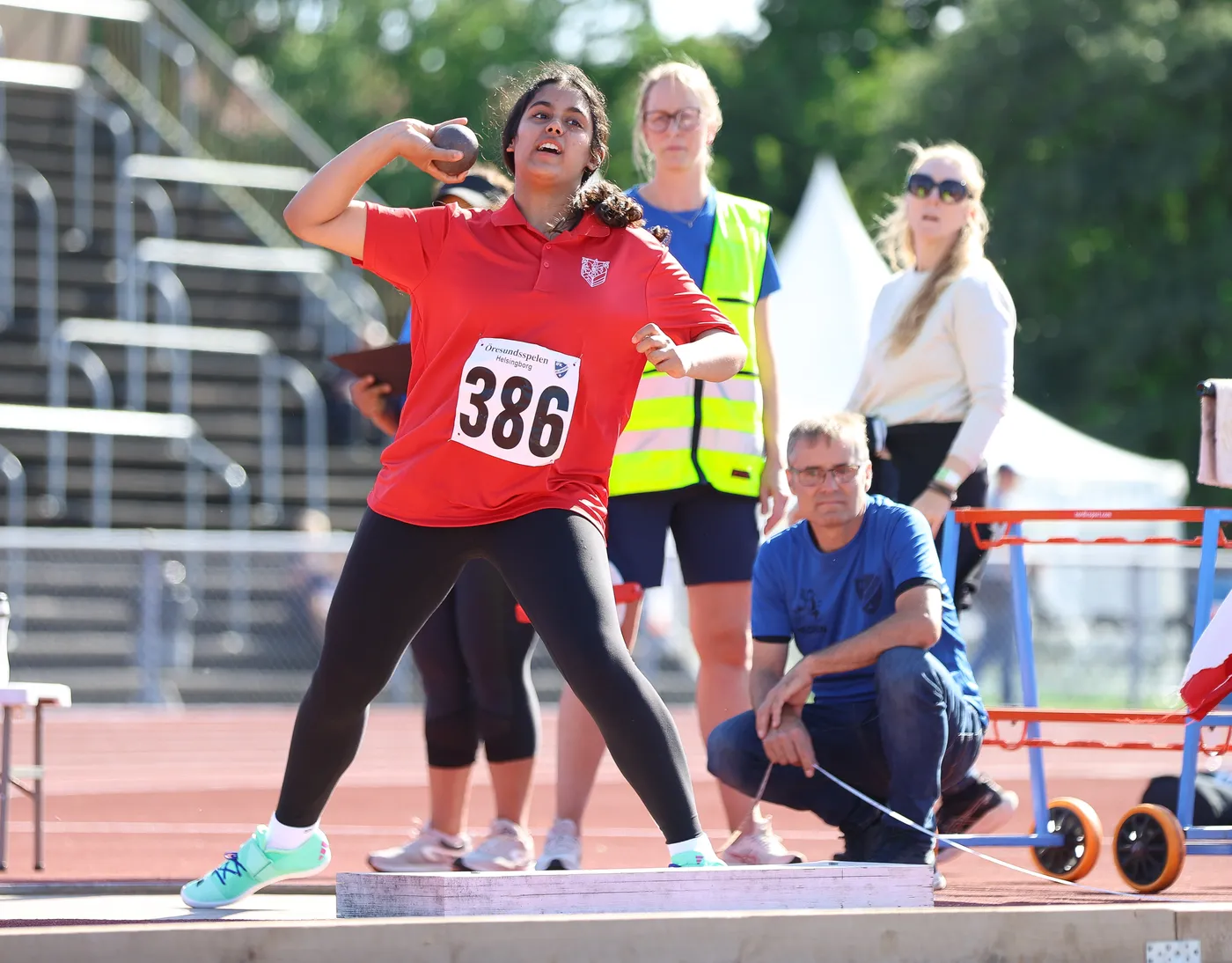 عداءة ترتدي قميصًا أحمرًا مع رقم 386، تستعد لرمي الكرة الحديدية في مسابقة رياضية. خلفها يُمكن رؤية عدد من الأشخاص، بما في ذلك بعض المدربين والمشجعين، وتبدو الأجواء حماسية مع وجود مضمار رياضي في الخلفية.