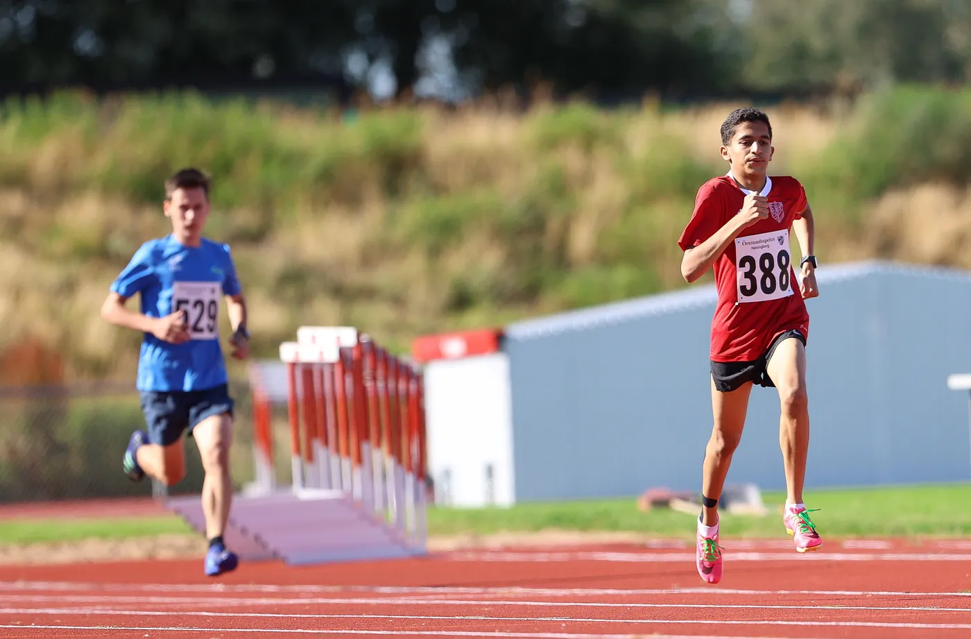 عداء يرتدي قميصاً أحمر يحمل الرقم 388 يجري بسرعة على المضمار, بينما يوجد عداء آخر يرتدي قميصاً أزرق يحمل الرقم 529 خلفه. تظهر حواجز رياضية في الخلفية مع منظر طبيعي أخضر.