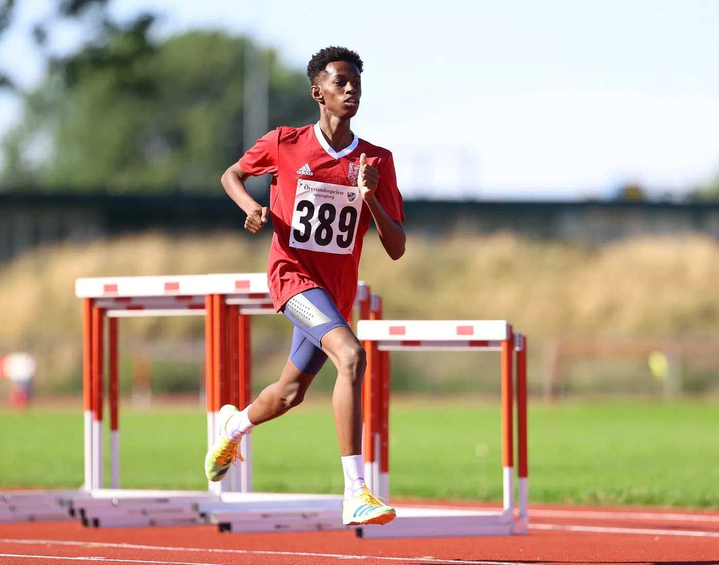 عداء شاب يرتدي زيًا رياضيًا أحمر يحمل الرقم 389، ويقوم بالجري على مضمار مخصص لسباقات الحواجز. تظهر الحواجز في الخلفية والسماء صافية، مما يشير إلى يوم مشمس.