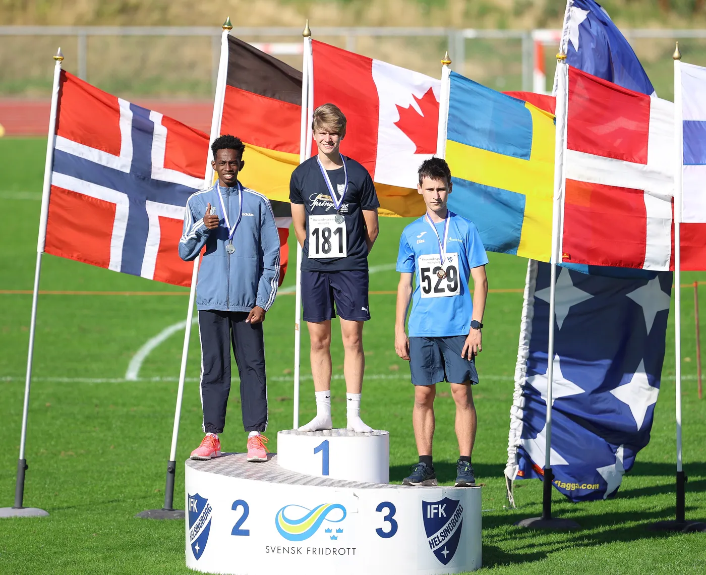 يقف ثلاثة رياضيين على منصة التتويج، حيث يرتدي الثنائي الأول والثاني ميداليات ويتبادلون الابتسامات، بينما يحمل الثالث علامة رقمية. خلفهم ترفرف أعلام متعددة لدول مختلفة، مثل النرويج وكندا وألمانيا والسويد. الجو يبدو مشمسًا وصيفياً في ملعب رياضي.