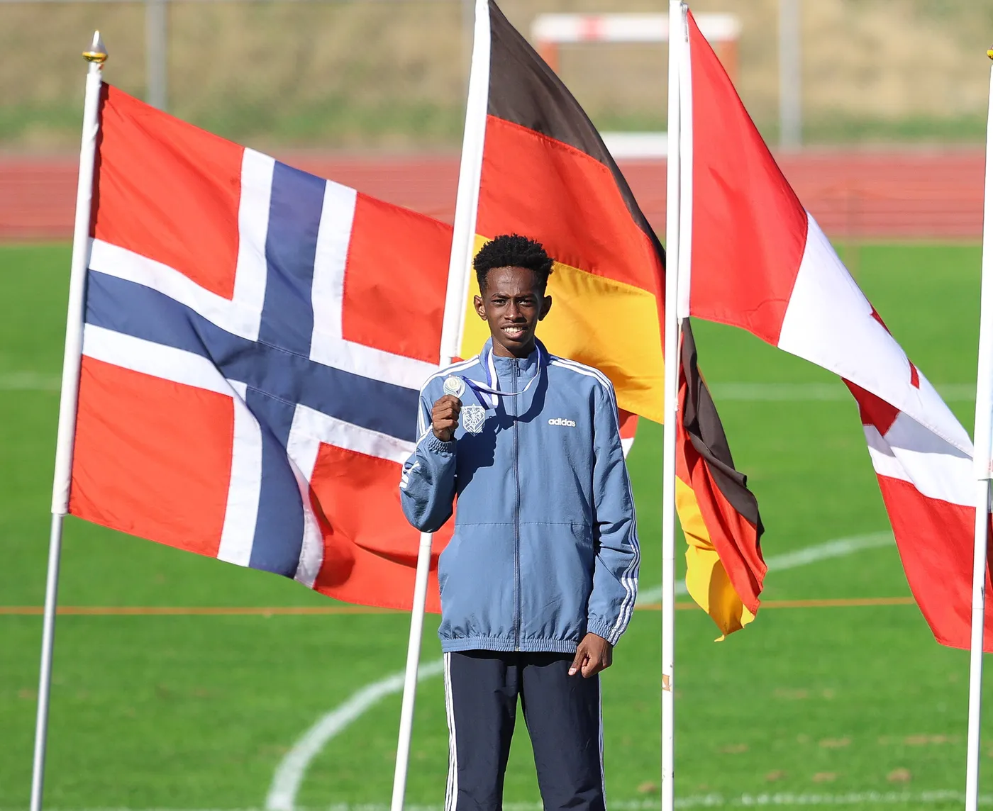 شاب يحمل ميدالية فضية، يقف مبتسمًا أمام أعلام متعددة لألمانيا والنرويج ودول أخرى، مع خلفية خضراء تمثل ملعب رياضي. توحي تعابير وجهه بالفرح والاعتزاز.