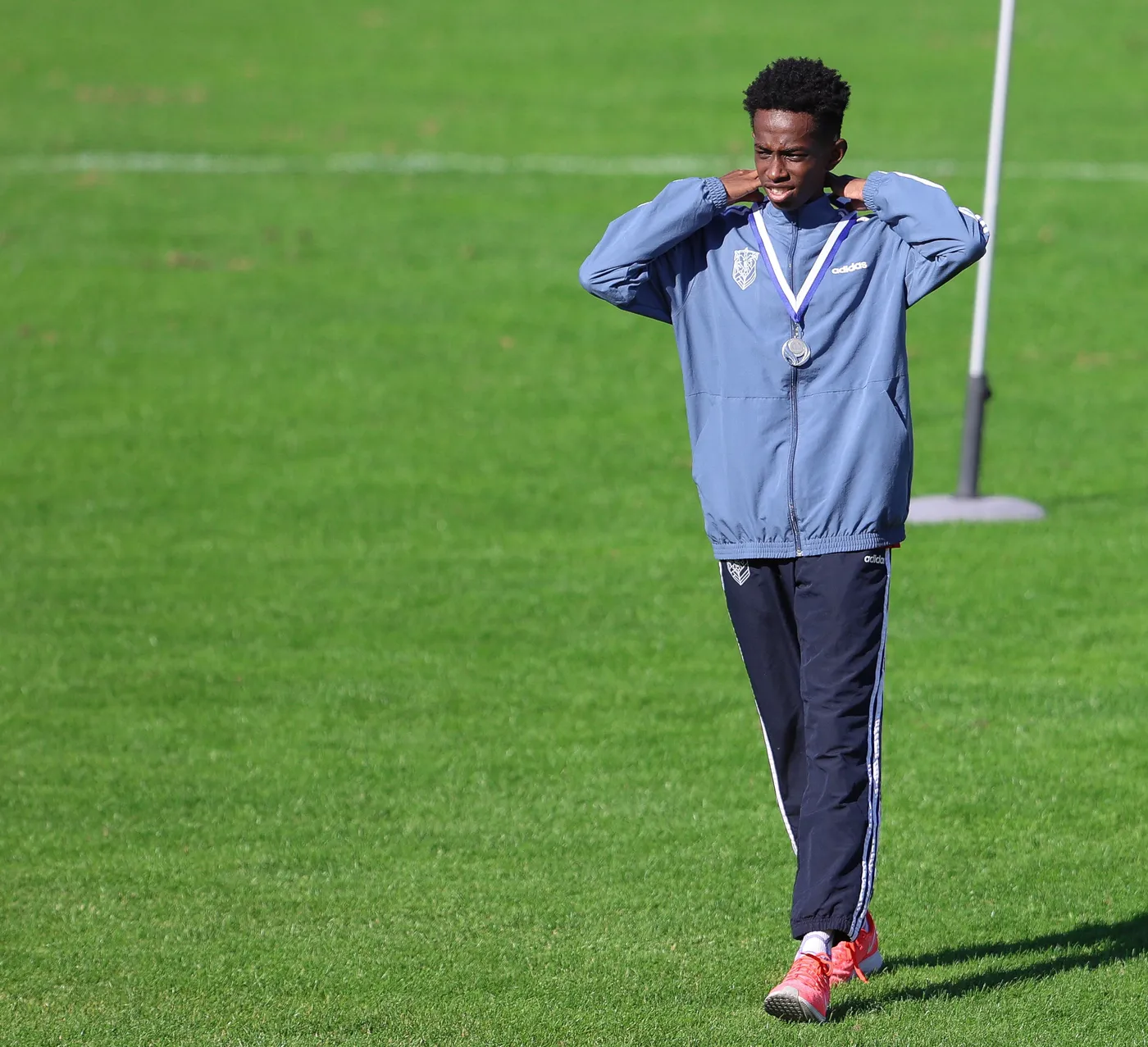 شاب يرتدي جاكيت رياضي وزوجًا من السراويل الرياضية، يحمل ميدالية فضية حول رقبته، يمشي على العشب الأخضر وهو يظهر ملامح الفخر.